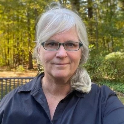A woman with gray hair and glasses taking a selfie outdoors in a wooded area.