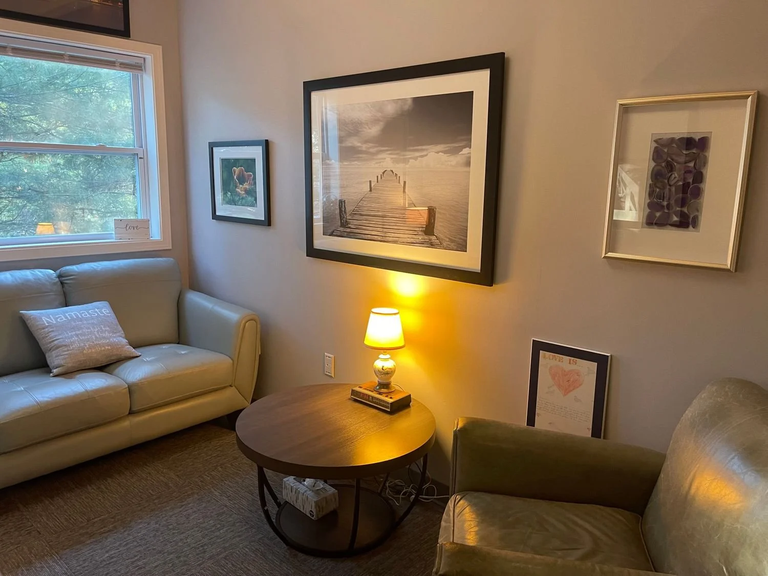 Living room with a beige sofa, a wooden side table with a lamp, a green armchair, and framed artwork on the wall. Window with trees outside.
