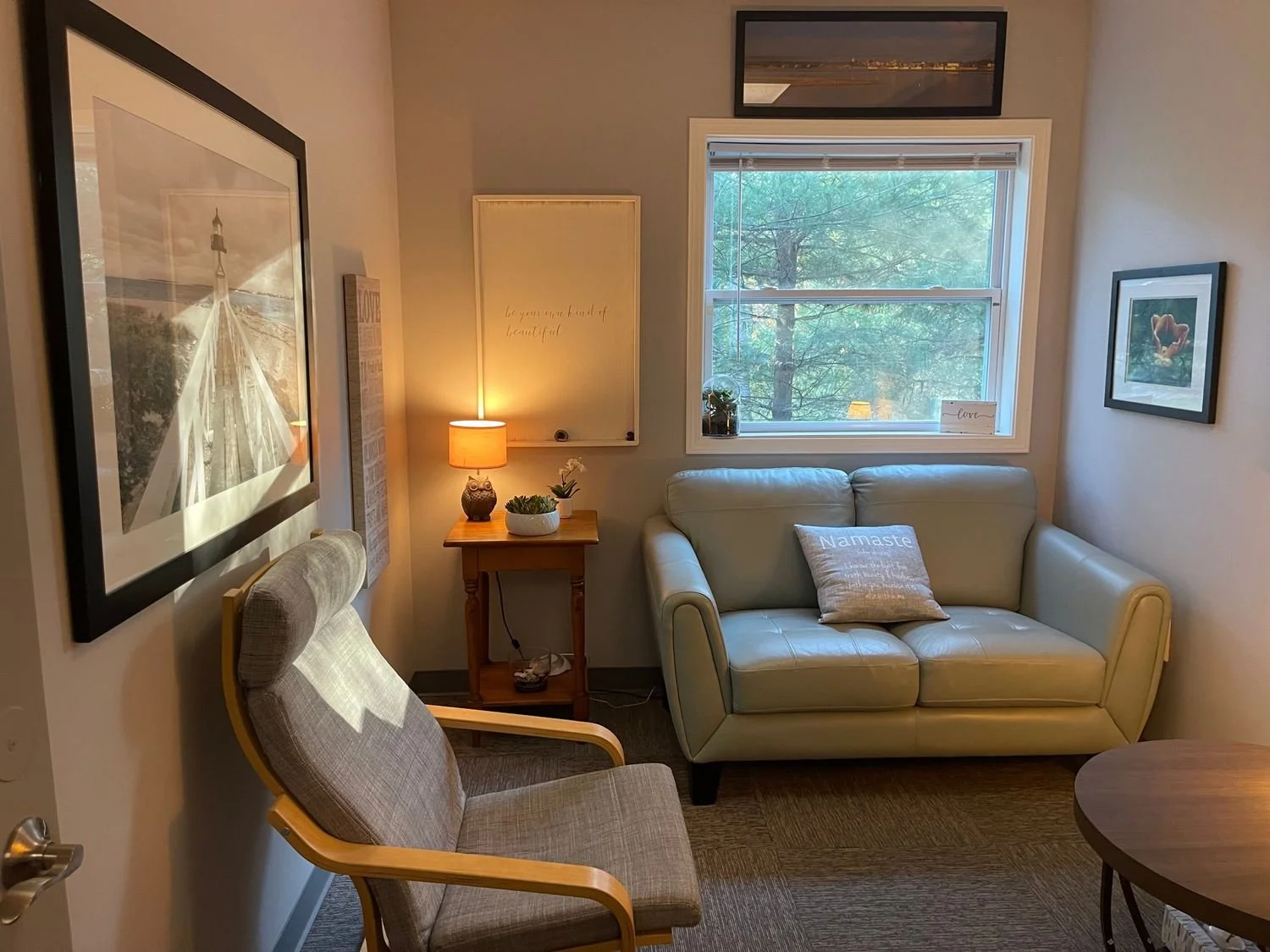 A cozy living room with a beige couch, a grey armchair, a wooden side table with a lamp and plants, framed artwork on the walls, a window showing trees outside, and decorative pillows.