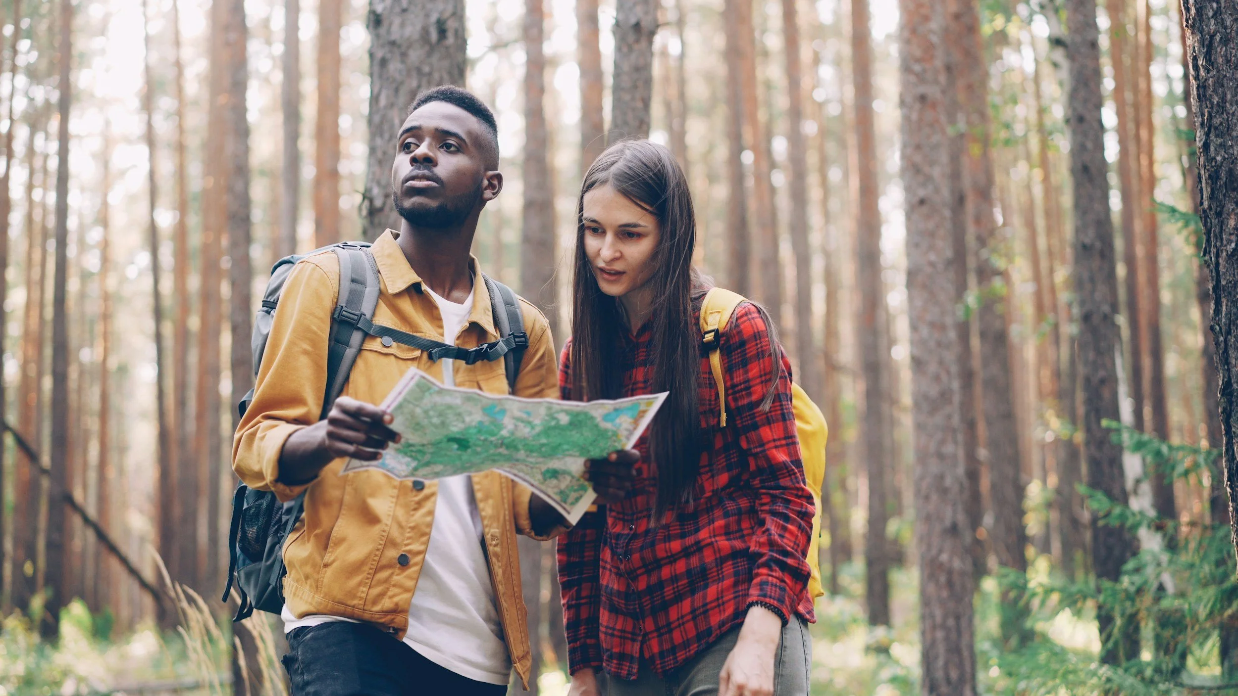Two hikers, a man and a woman, standing in a forest, looking at a map together with tall trees in the background.