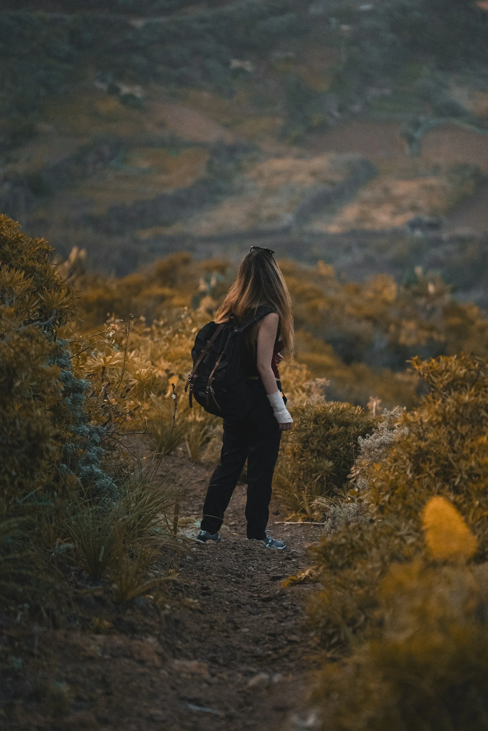 A person with long hair, wearing a tank top and black pants, carries a backpack and walks along a trail surrounded by yellowish bushes, with mountain landscape in the background during dusk or early evening.