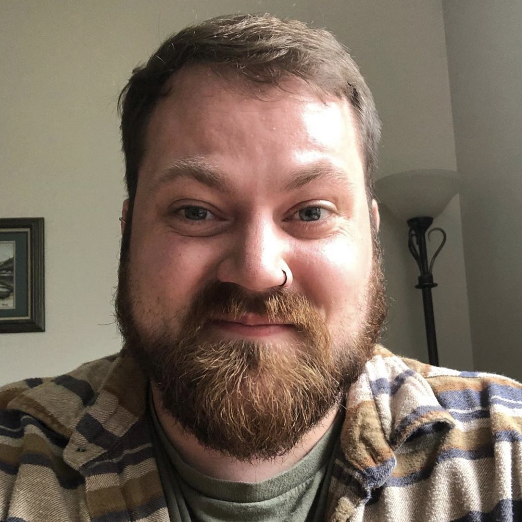 A man with a beard, mustache, and light skin in a plaid flannel shirt, smiling at the camera in a home setting with a wall lamp and framed picture in the background.
