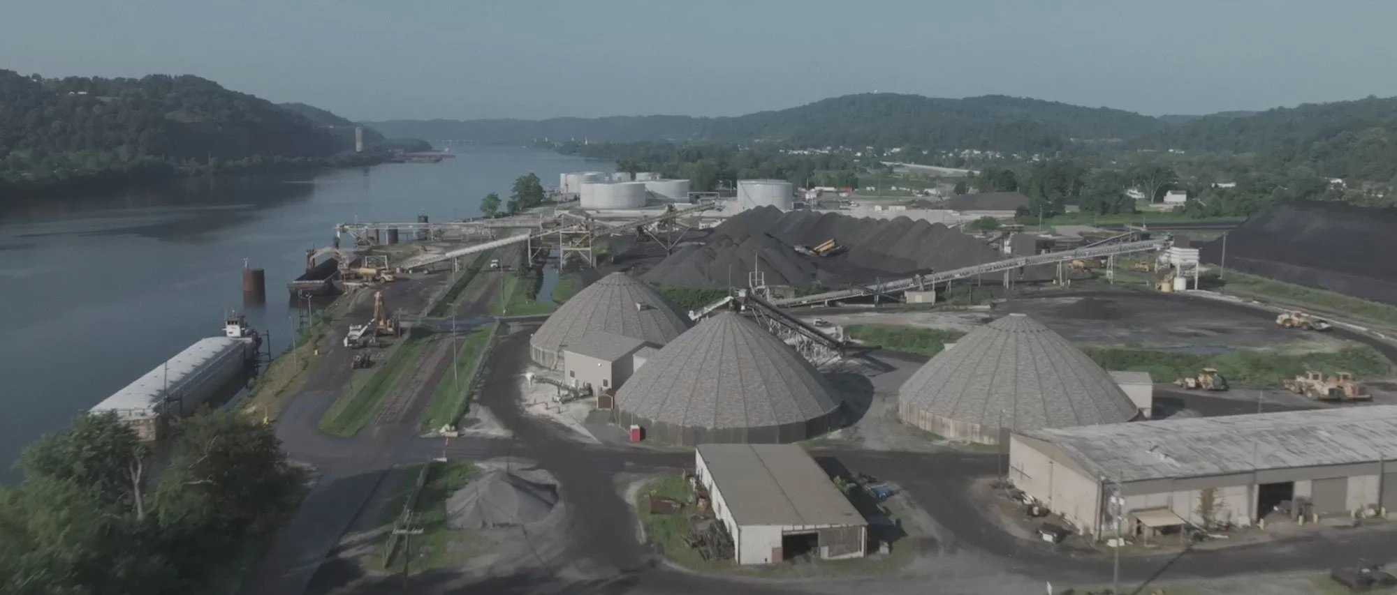 Pickands Mather Supply Chain Consulting | An industrial facility with large, cone-shaped storage structures near a body of water, surrounded by machinery, conveyor belts, and vehicles, with hills and greenery in the background.