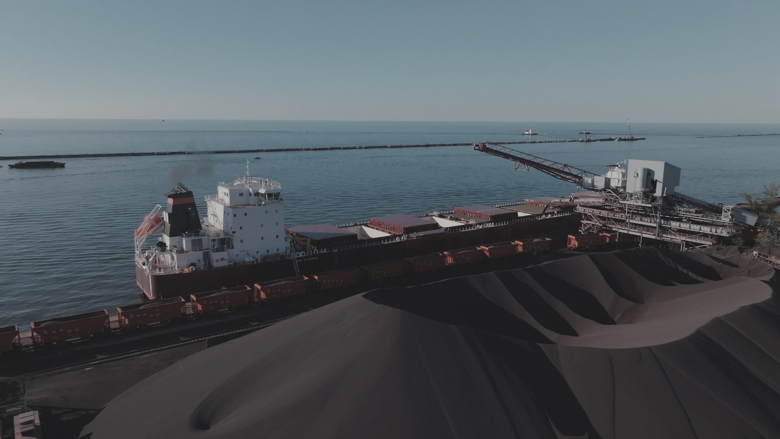 Pickands Mather Freight Forwarding | A large cargo ship with a black and white hull sailing near a coal stockpile by the water, with a long breakwater and several smaller boats in the distance on a clear day.