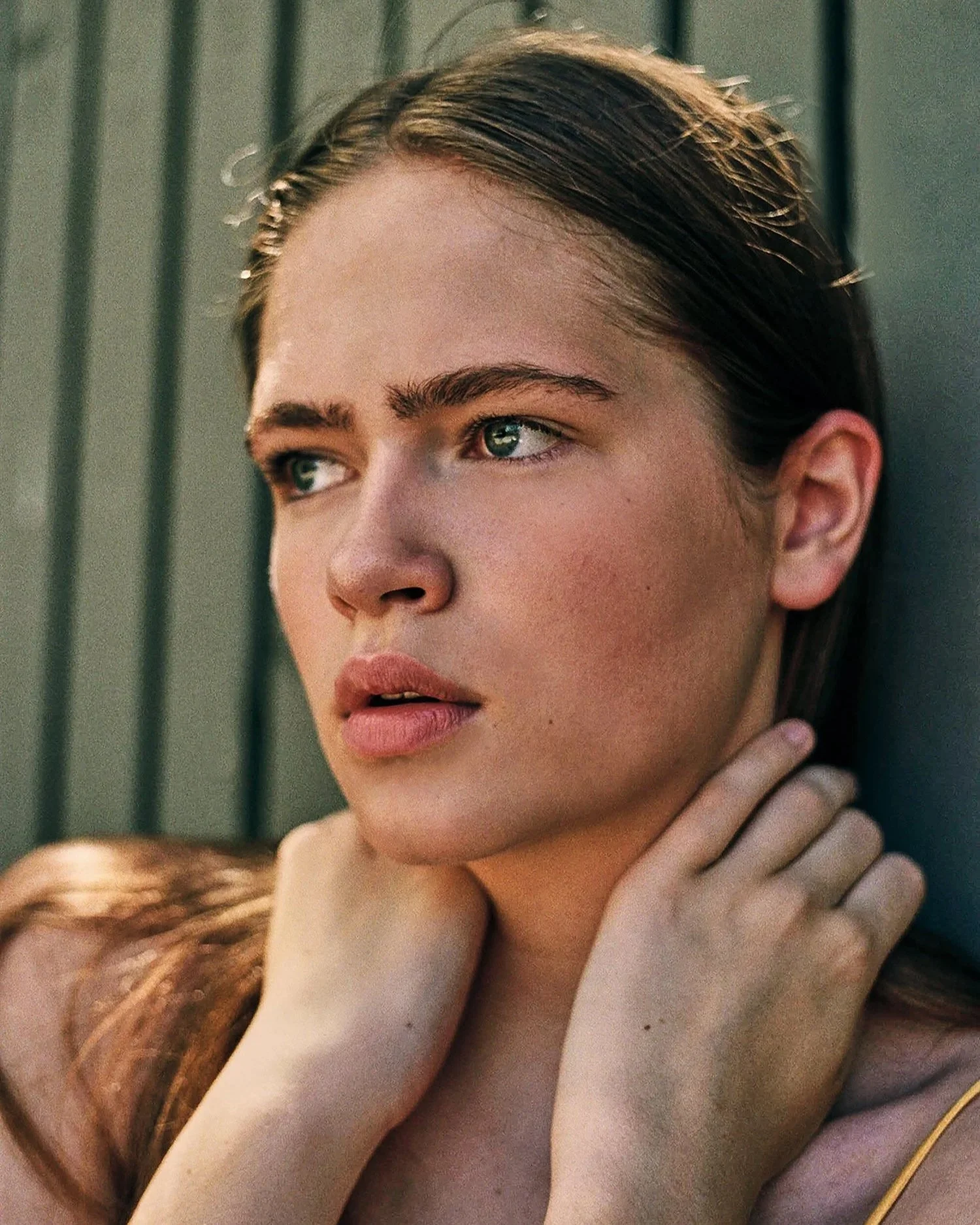Close-up of a young woman with blue eyes and brown hair, standing against a gray wall, looking thoughtfully to the side with her hand resting on her neck.