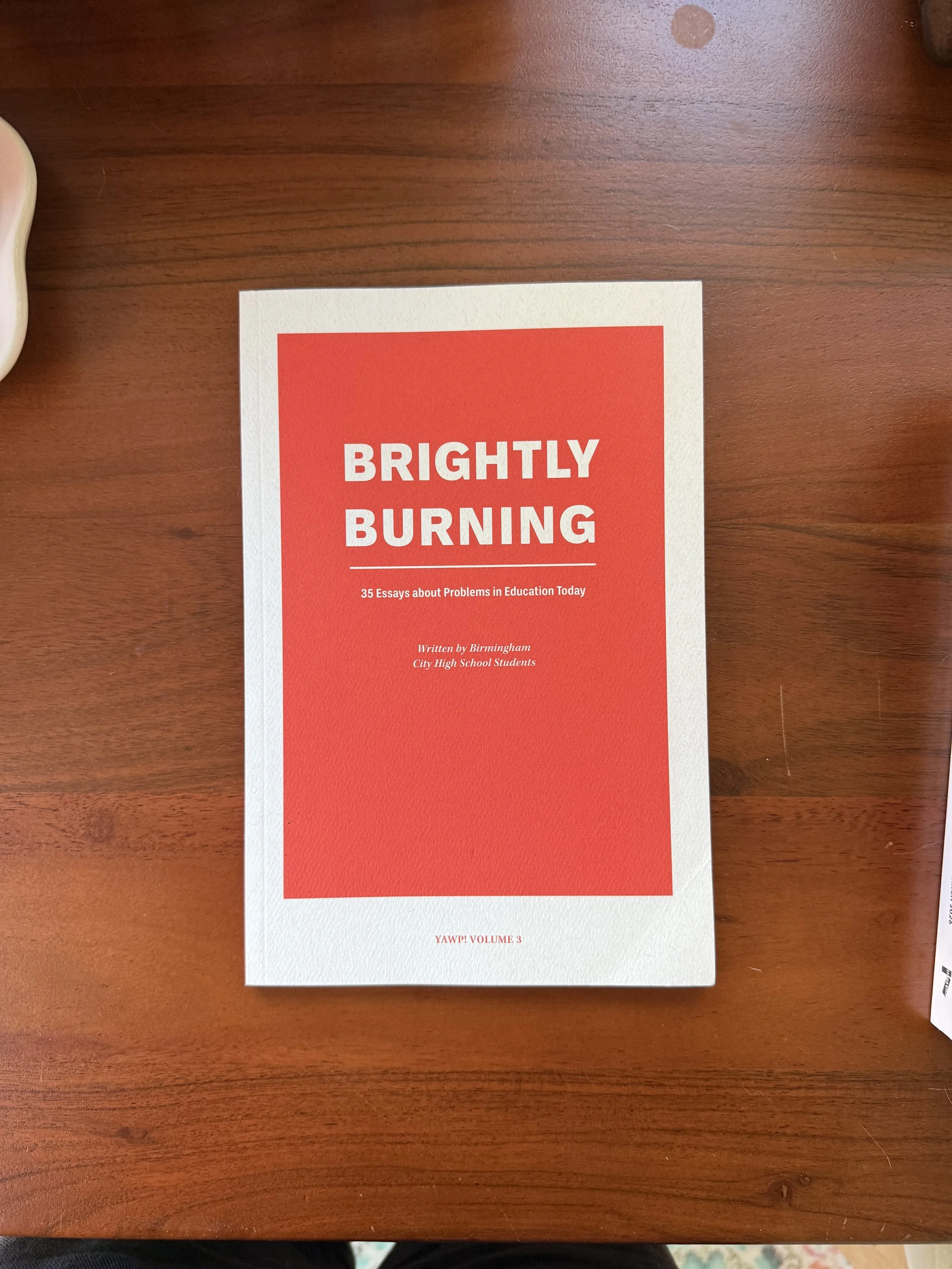 A book titled "Brightly Burning" with a red cover, placed on a wooden table.