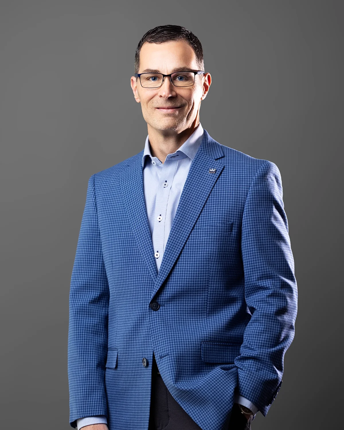 Portrait of Neal Juern in a blue checkered blazer and light blue button-up shirt, wearing glasses, standing against a plain gray background.
