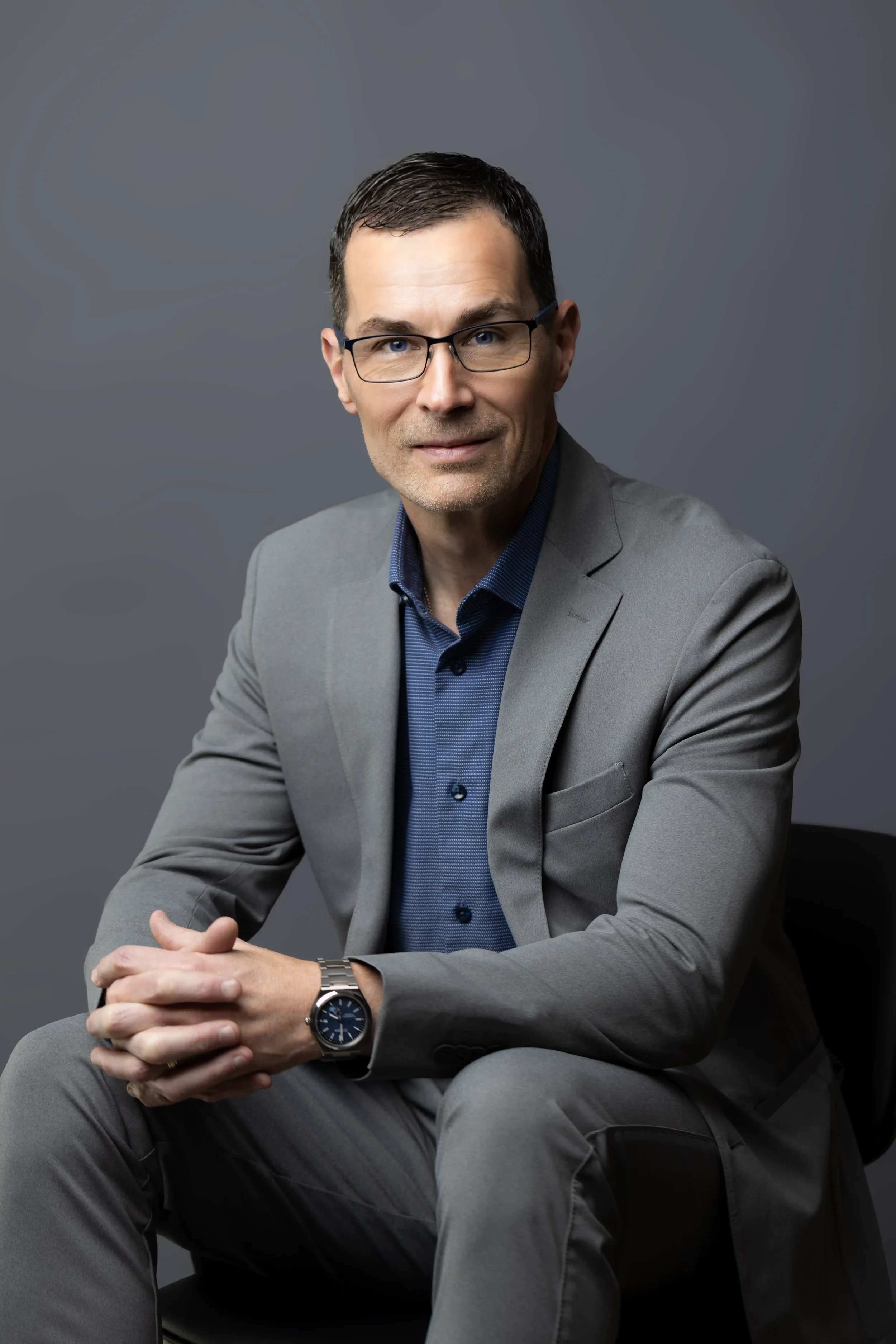Portrait of Neal Juern wearing a gray suit and blue shirt, sitting with hands clasped, against a plain gray background.