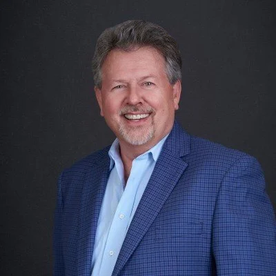 A middle-aged man with gray hair and a beard, smiling, wearing a blue suit jacket and a light blue dress shirt, against a dark background.