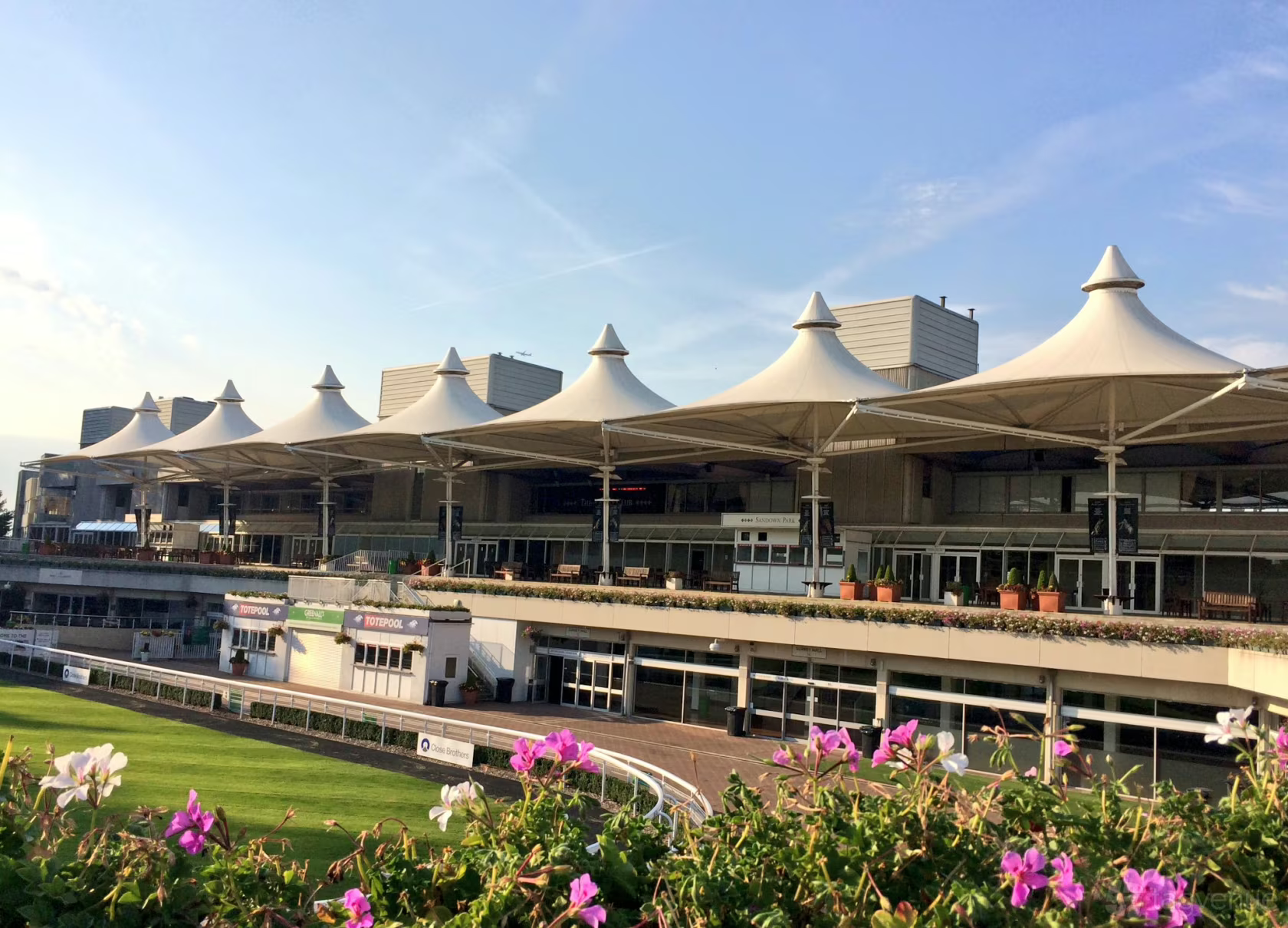 A sunny afternoon view of the grandstand at Sandown Park Racecourse, characterized by a prominent row of white, cone-shaped tensile fabric canopies.