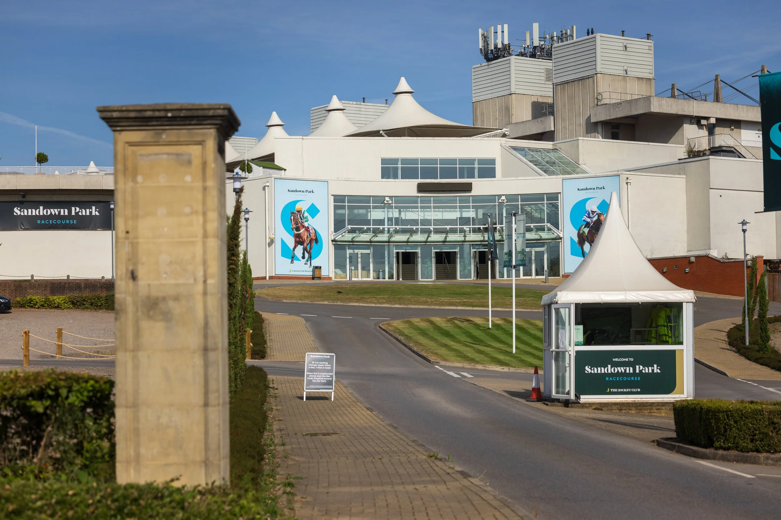 Exterior view of Sandown Park Racecourse with banners, tents, and pathways, under a clear blue sky.