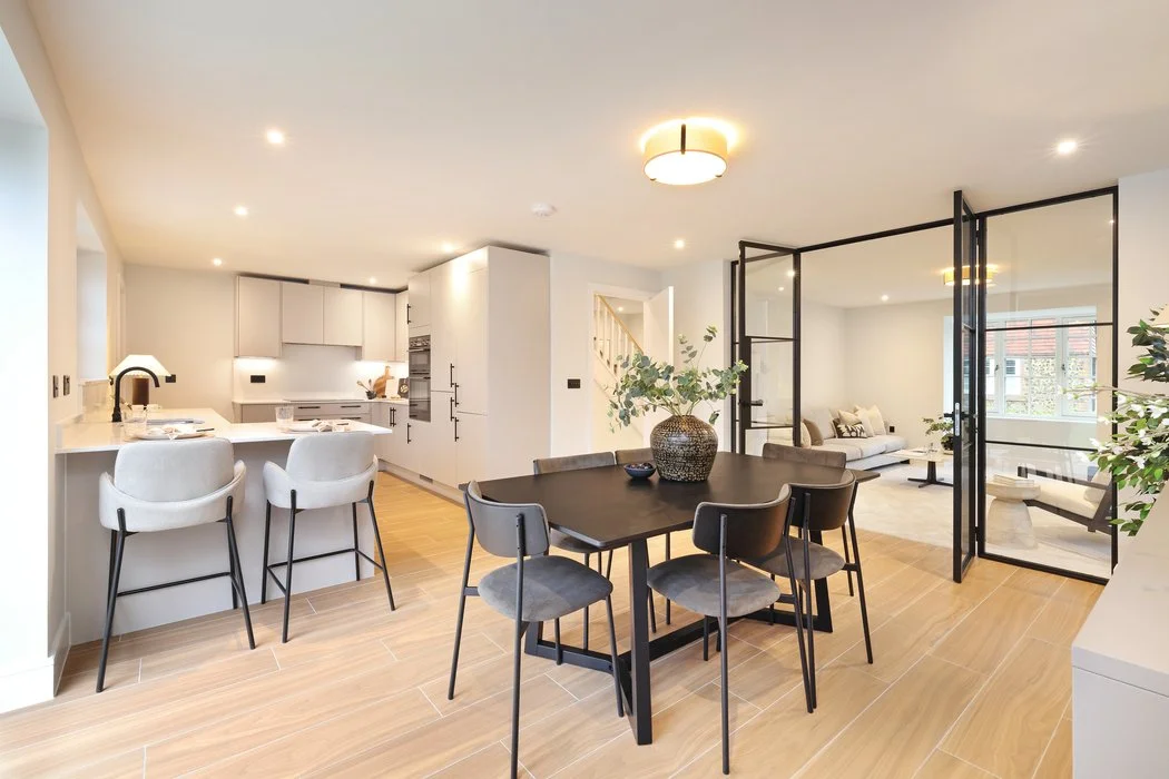 A bright, contemporary open-plan home interior featuring light wood flooring. In the centre is a dark rectangular dining table surrounded by six grey chairs. To the left is a sleek white kitchen with built in appliances and an island breakfast bar.