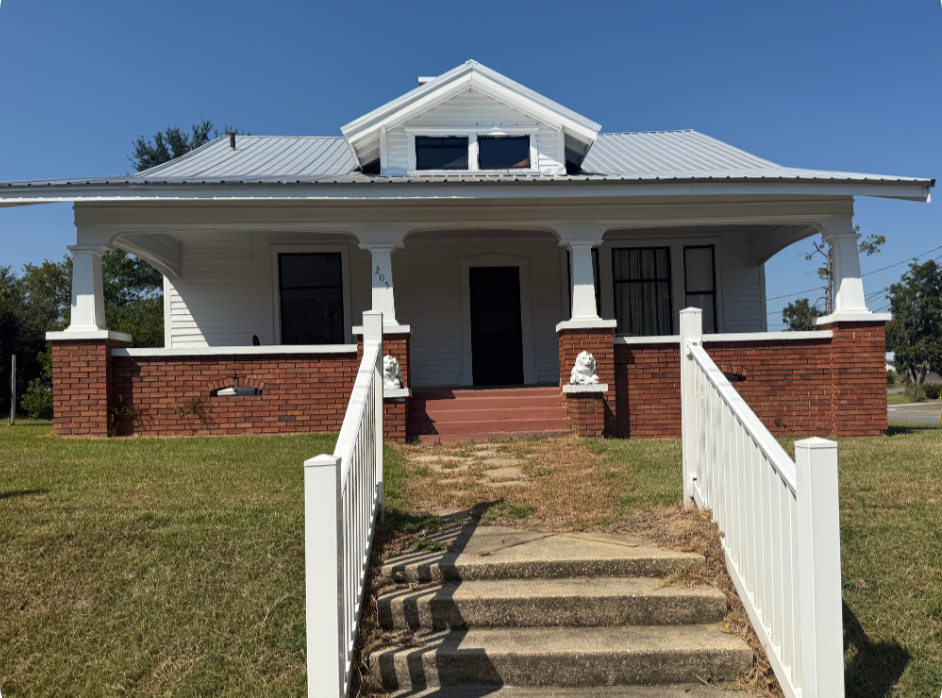 White house with red brick foundation, front porch, and stairs, surrounded by a grassy yard, under a blue sky.
