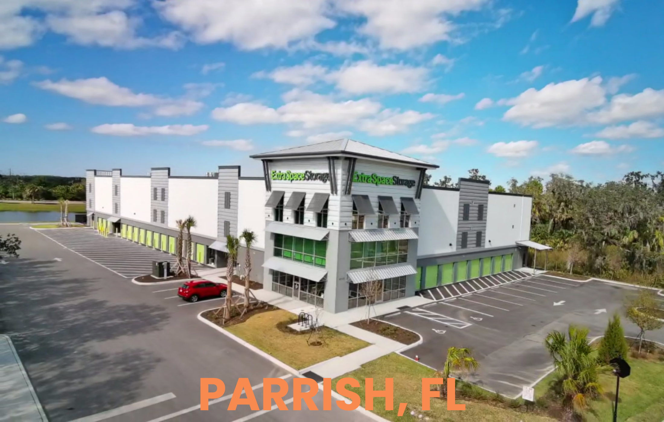 An ariel, drone photo of a modern, three-story storage building with green accents, palm trees, and a blue sky with clouds, labeled PARRISH, FL.