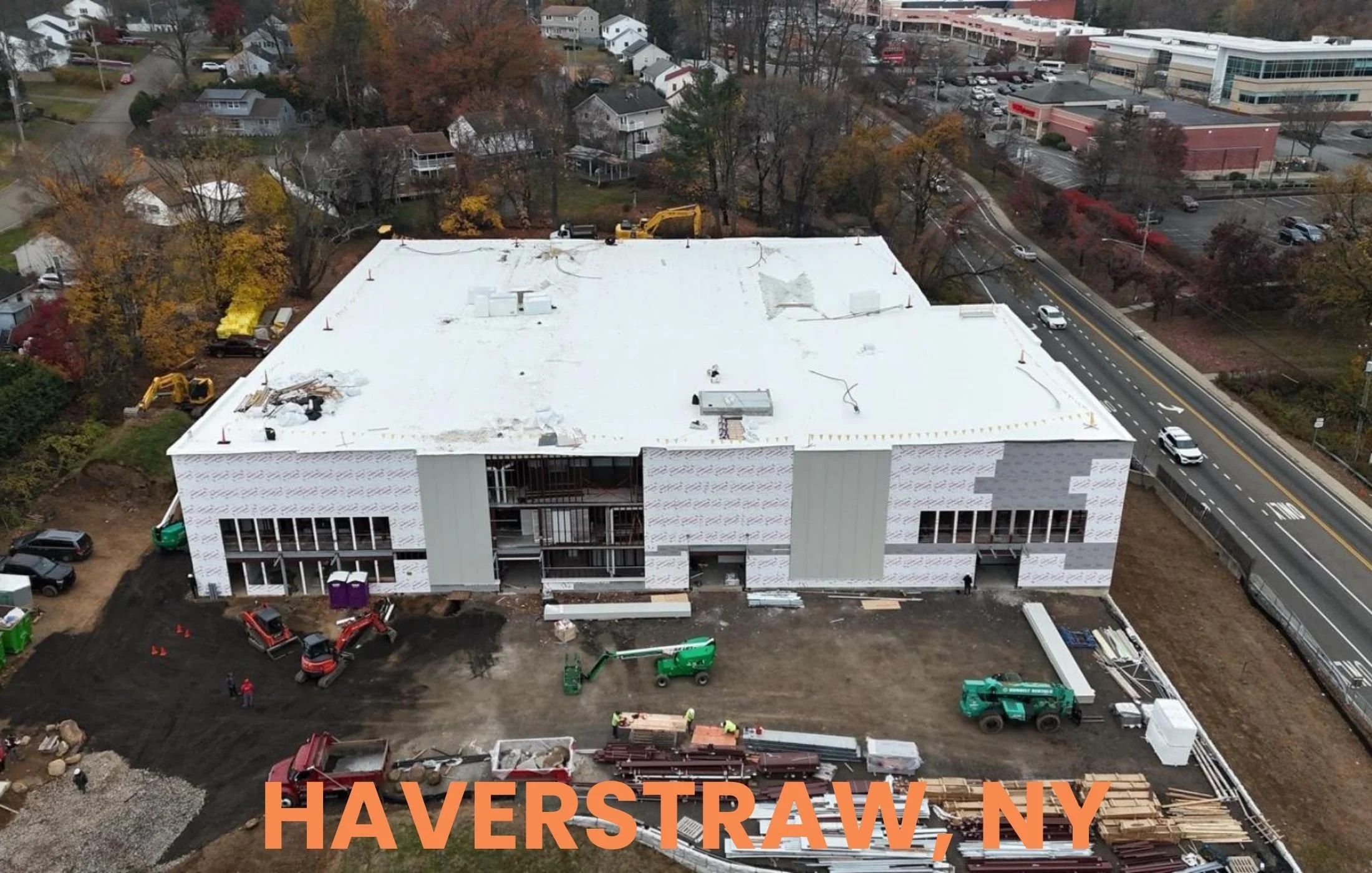 Aerial view of a building under construction in Haverstraw, NY. The building has a white roof and partially completed exterior walls. Construction equipment and workers are present on the site, with trees and nearby residential and commercial buildin