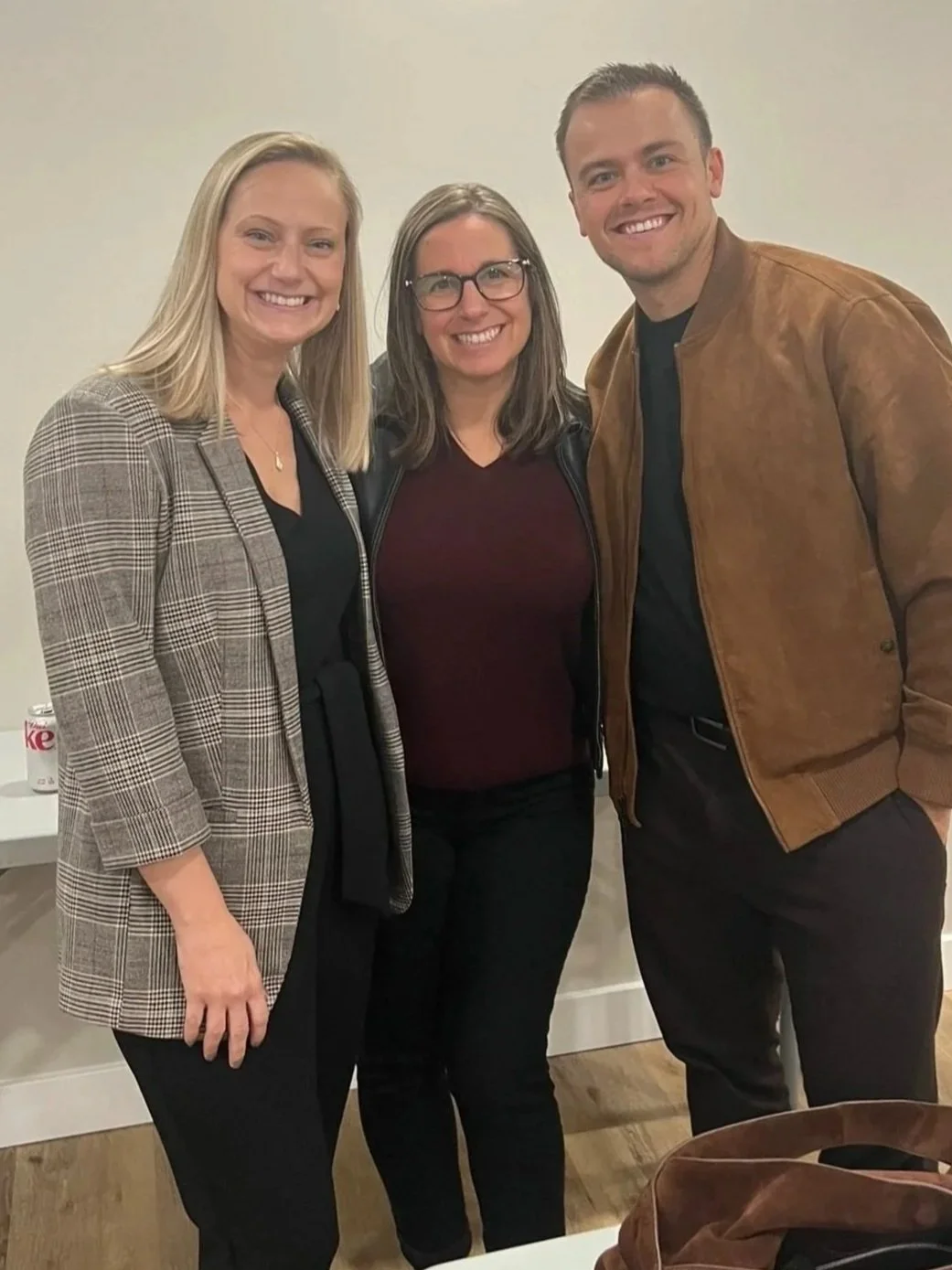Three smiling adults, two women and one man, standing closely together against a plain off-white wall. The woman on the left has blonde hair and is wearing a checked blazer. The woman in the middle has shoulder-length brown hair, glasses, and a black jacket. The man on the right has short brown hair and is wearing a brown leather jacket.