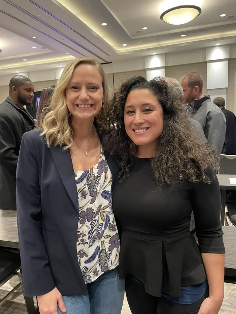Two women smiling and posing for a photo in a conference room or event space, with other people in the background.