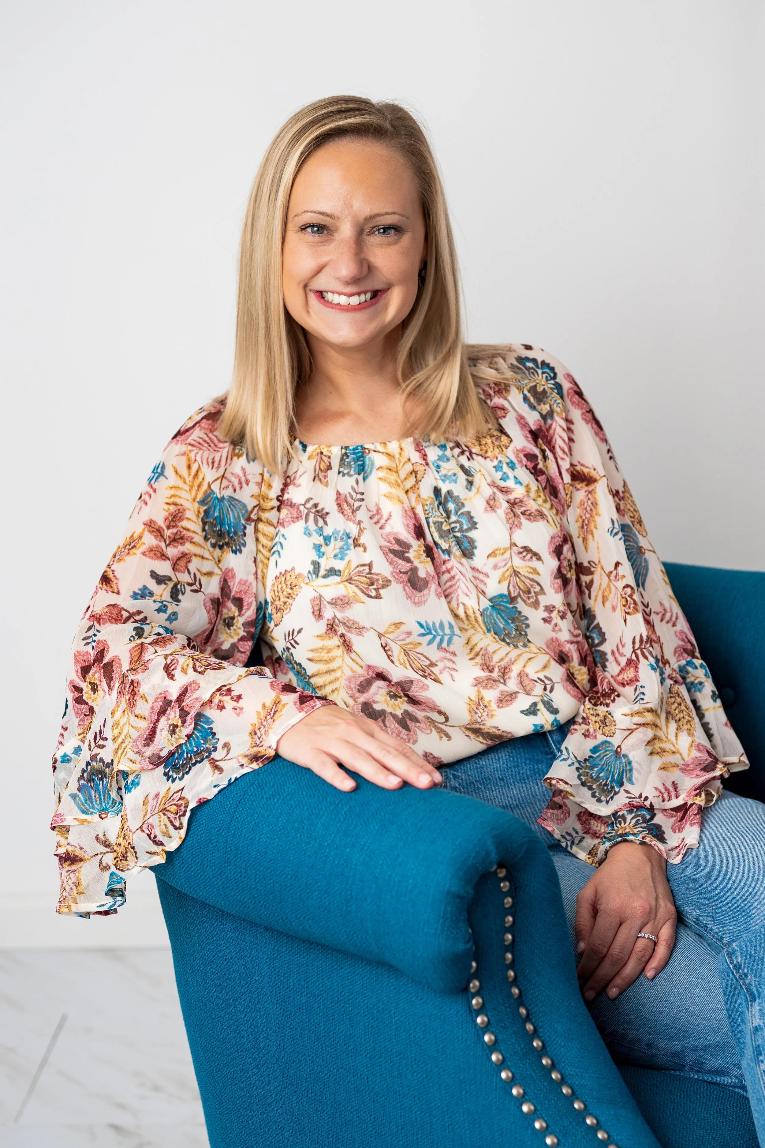 A woman with blonde hair smiling and sitting on a blue armchair, wearing a floral blouse and jeans, against a plain light background.