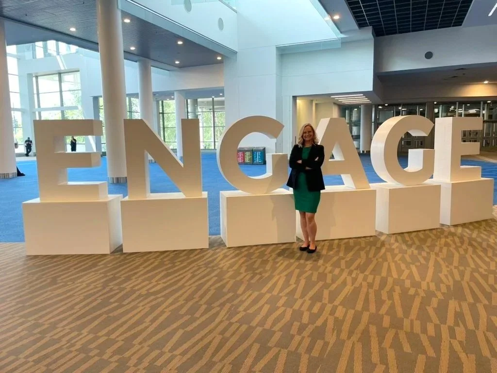 A woman standing next to large 3D letters spelling 'ENGAGE' inside a modern building with glass walls and blue carpet.