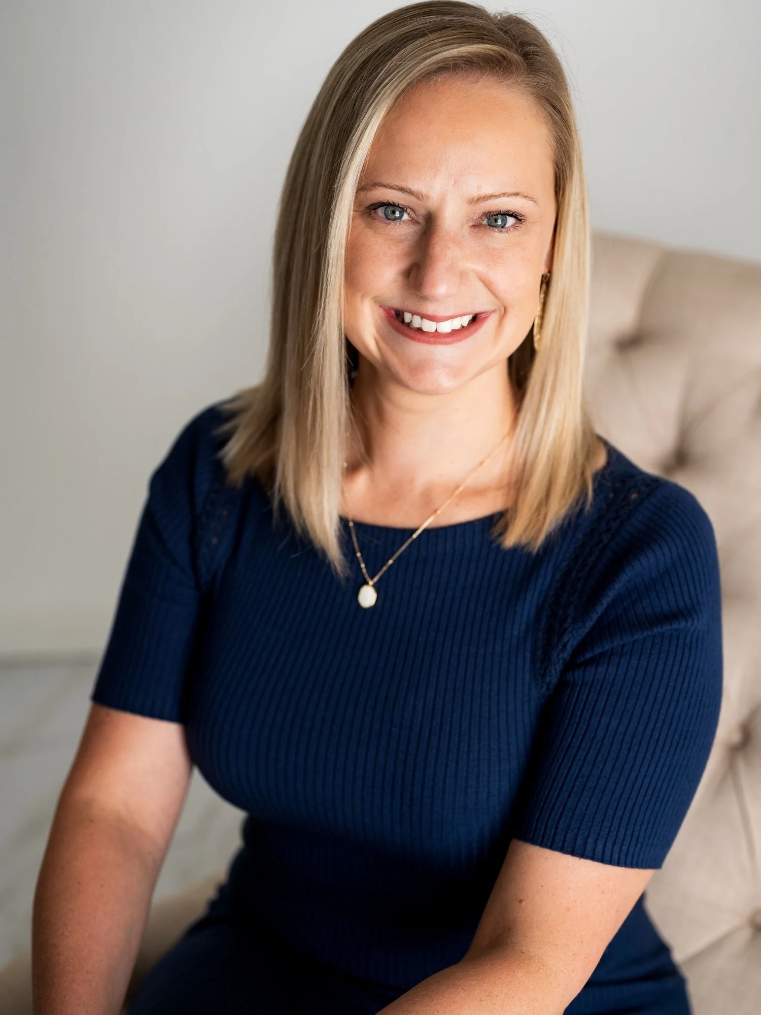 A smiling woman with shoulder-length blonde hair, blue eyes, wearing a navy blue ribbed top and gold jewelry, sitting on a beige chair with a neutral background.