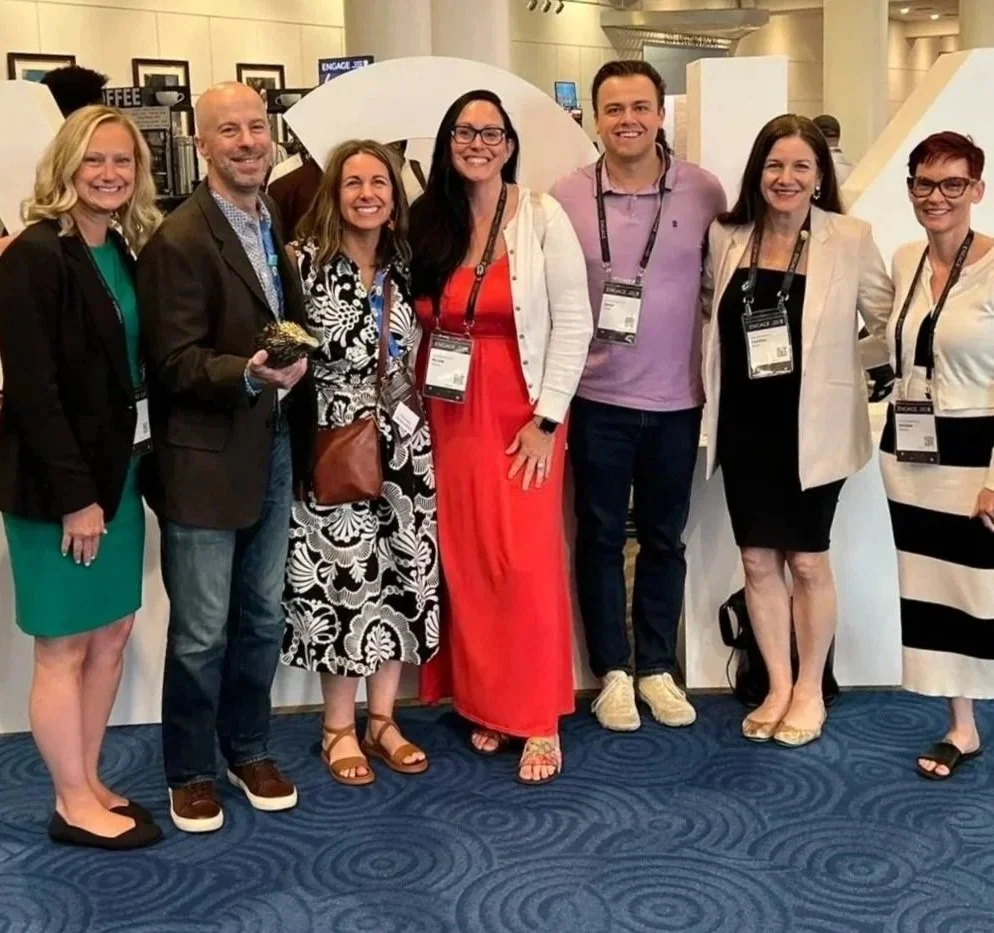 Group of seven people smiling and standing together at a conference or event, wearing name badges, in an indoor setting with booths and signs in the background.