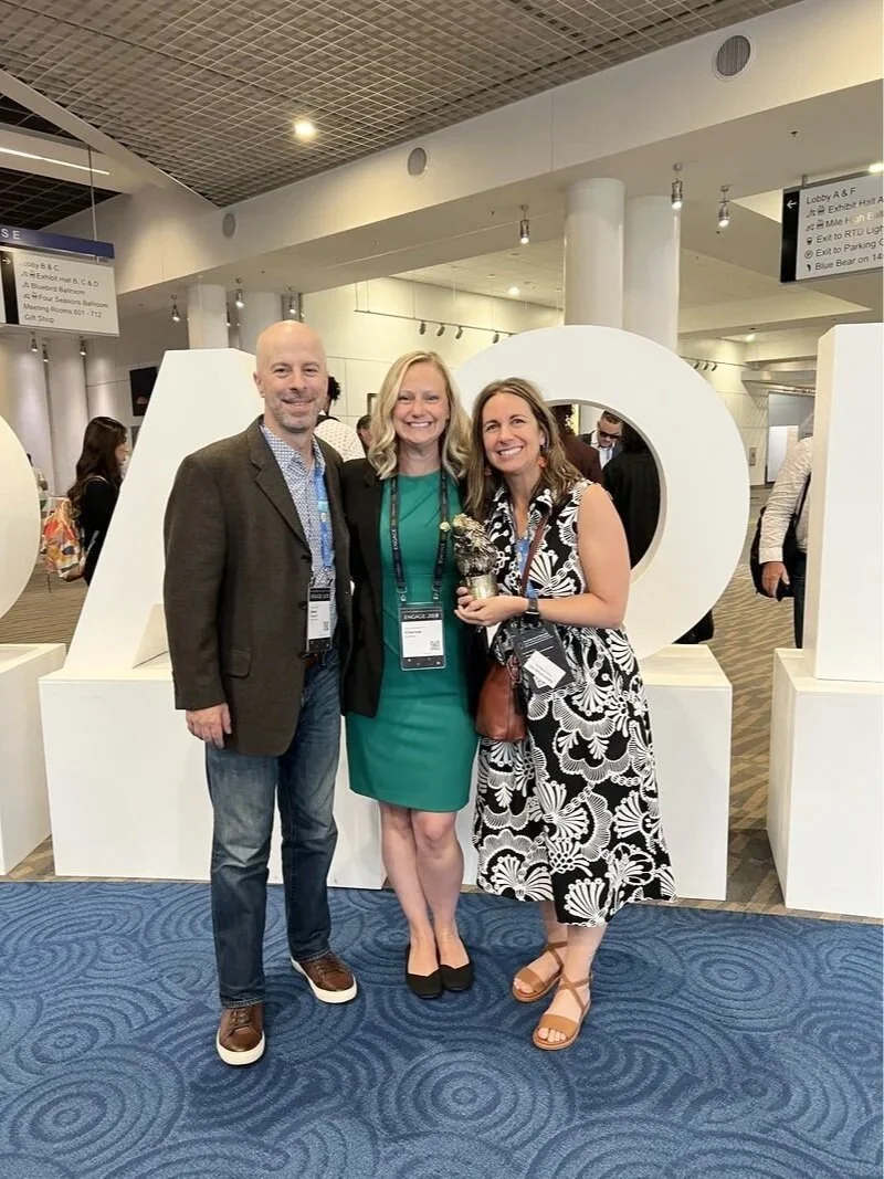 Three people standing together smiling at an indoor event, with large white decorative letters spelling 'LOVE' in the background, and conference badges around their necks.
