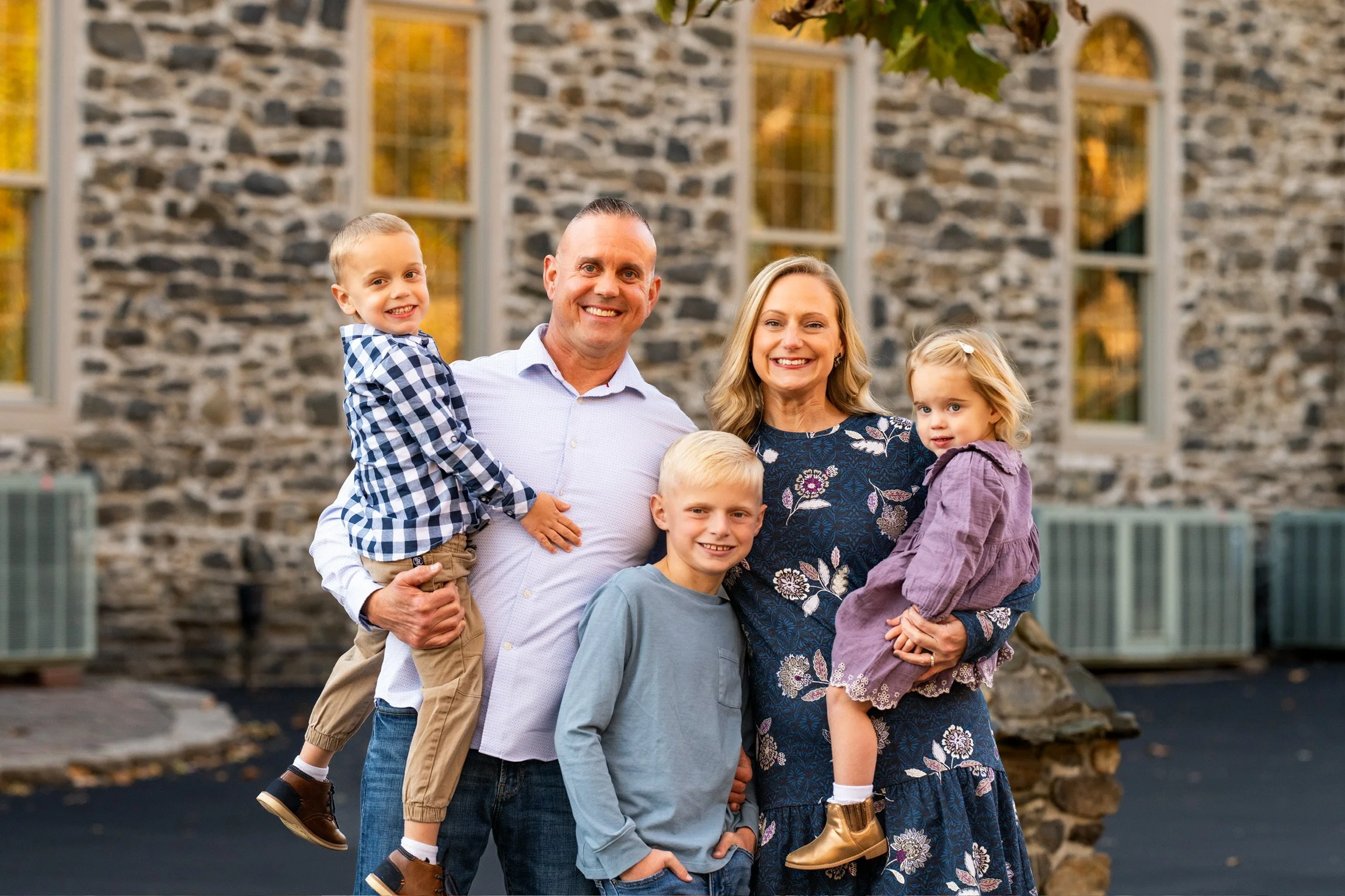 A smiling family of six posed outdoors in front of a stone building with large windows, during sunset. The family includes a man, woman, and their four children, two boys and two girls. The youngest boy is being held by the father, the older boy is standing in front of the mother, and the girls are being held by the parents.