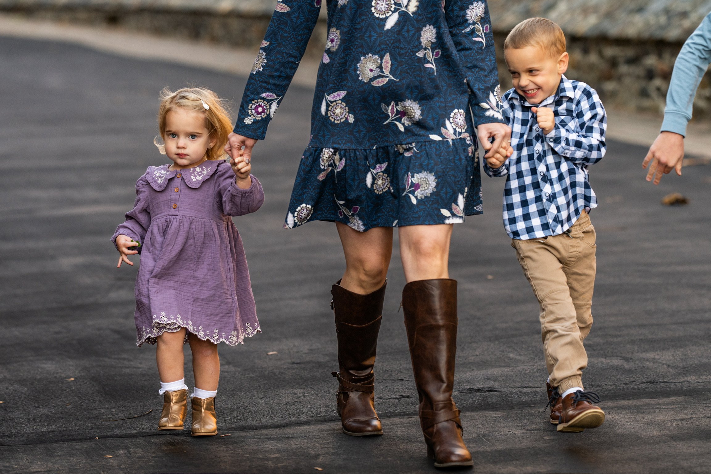 A woman walking on a paved road holding the hands of two children, a young girl in a purple dress and brown boots, and a boy in a blue plaid shirt and tan pants, with another person partially visible on the right.