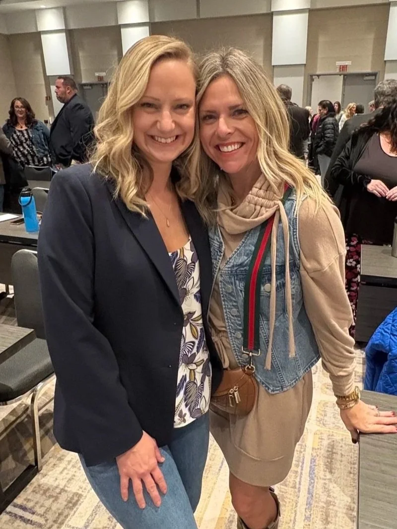 Two women smiling and posing together at an indoor event, with a crowd of people in the background.