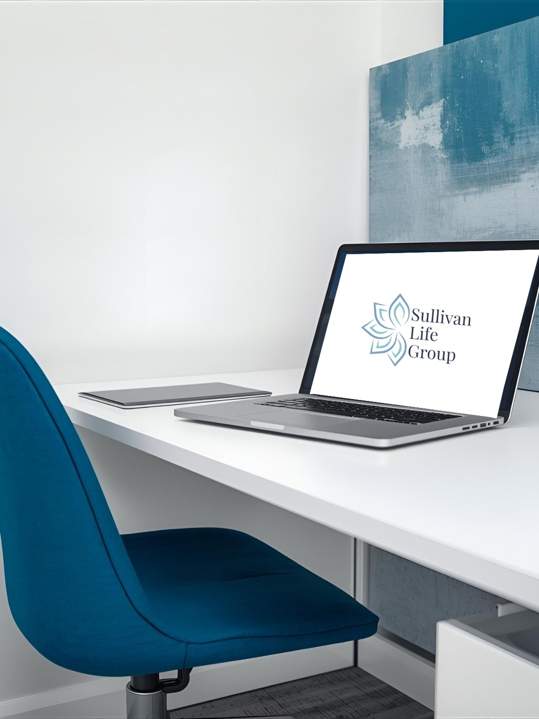 Office desk with a blue chair, a closed laptop, a tablet, and an open laptop displaying the Sullivan Life Group logo against a white wall with an abstract painting.