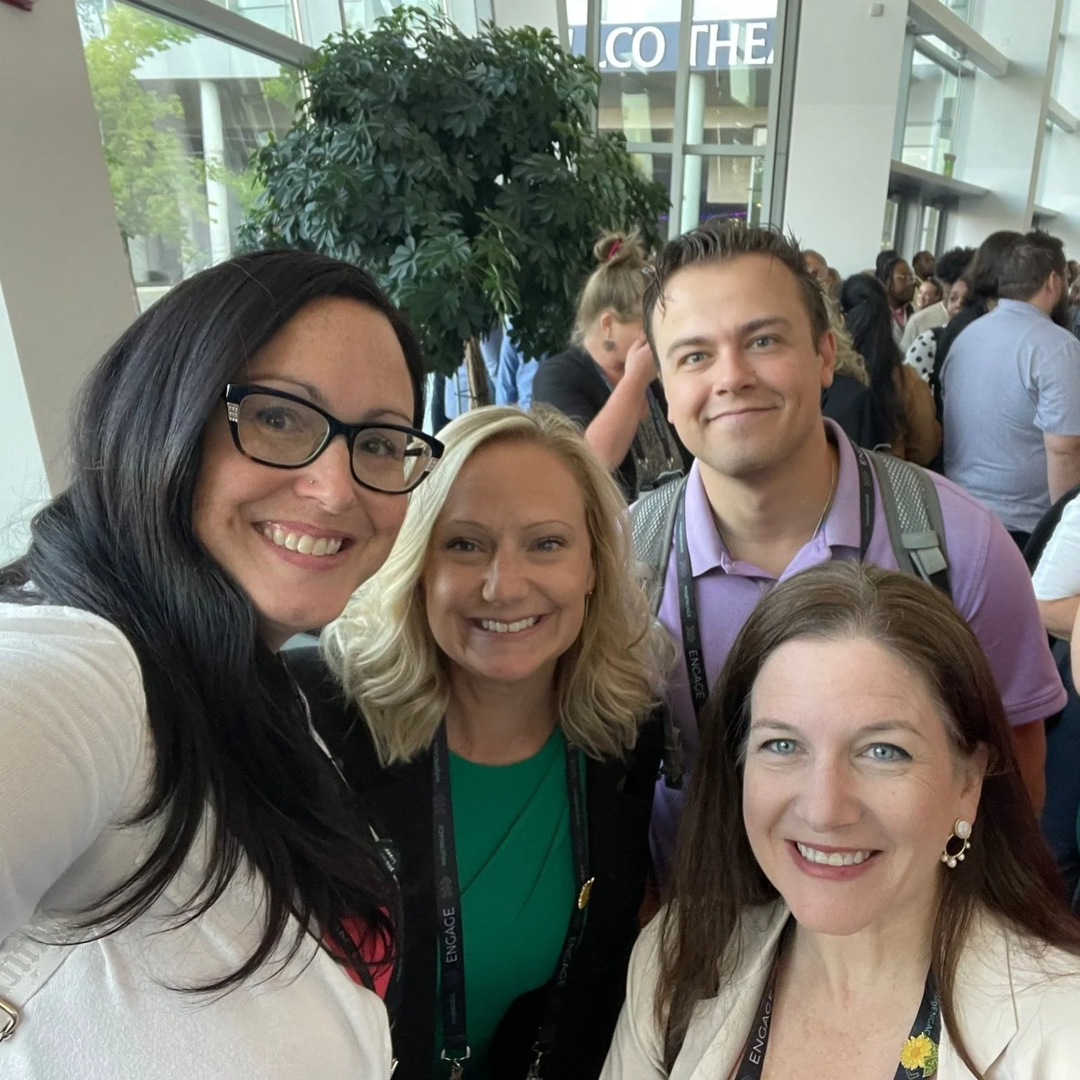Four women and one man taking a selfie at a conference or event, with a crowd of people behind them and glass windows revealing a tree outside.
