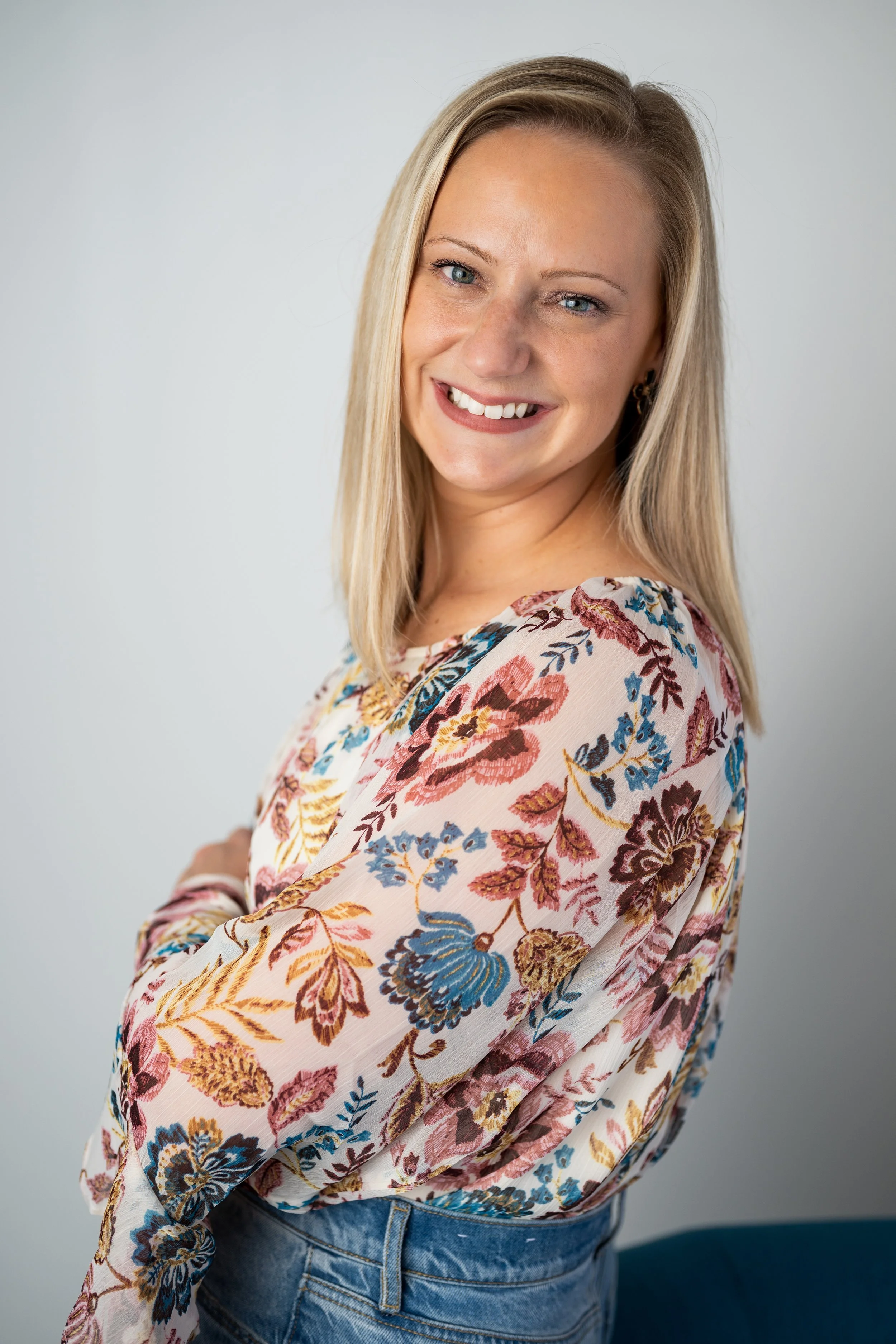 A young woman with blonde hair smiling, wearing a floral blouse with blue, pink, and yellow flowers, and blue jeans, standing against a plain light gray background.