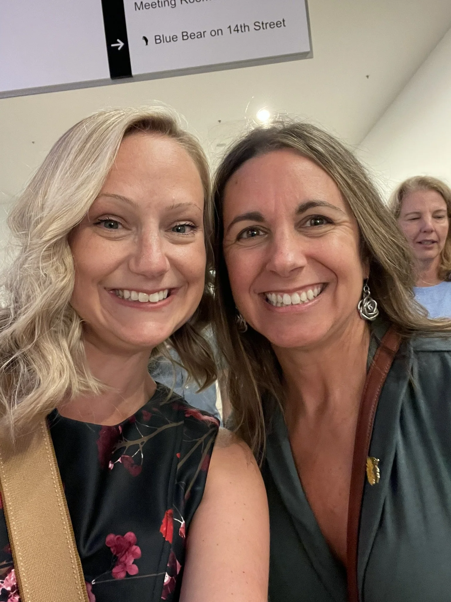 Two women smiling for a selfie in an indoor setting. One woman has blonde hair and is wearing a floral dress, and the other has brown hair and is wearing a dark jacket with earrings. In the background, another woman is visible, and there is a sign overhead that references a meeting room and a location on 14th Street.