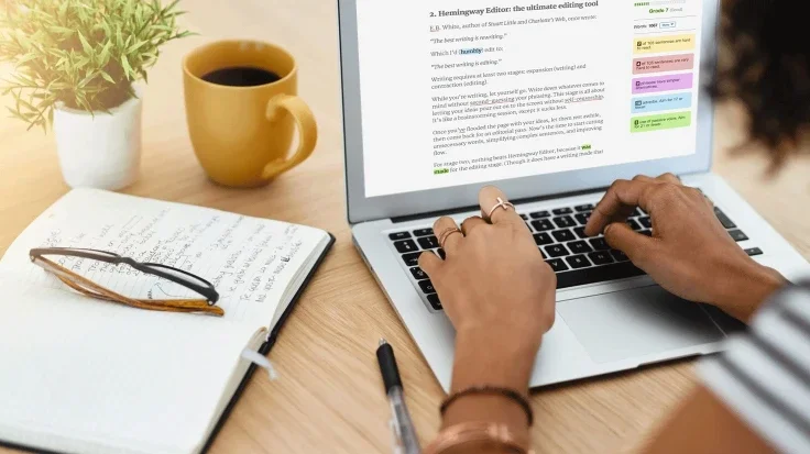 A person working on a laptop with an online document on the screen. There is an open notebook with a pen and a pair of glasses, a yellow coffee mug, and a potted plant on a wooden desk.