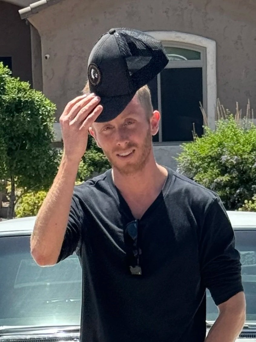 A young man with light brown hair and a beard is holding a black baseball cap on his head with his right hand. He is wearing a black t-shirt and sunglasses hanging from the shirt's collar, standing outdoors in front of a house with greenery.