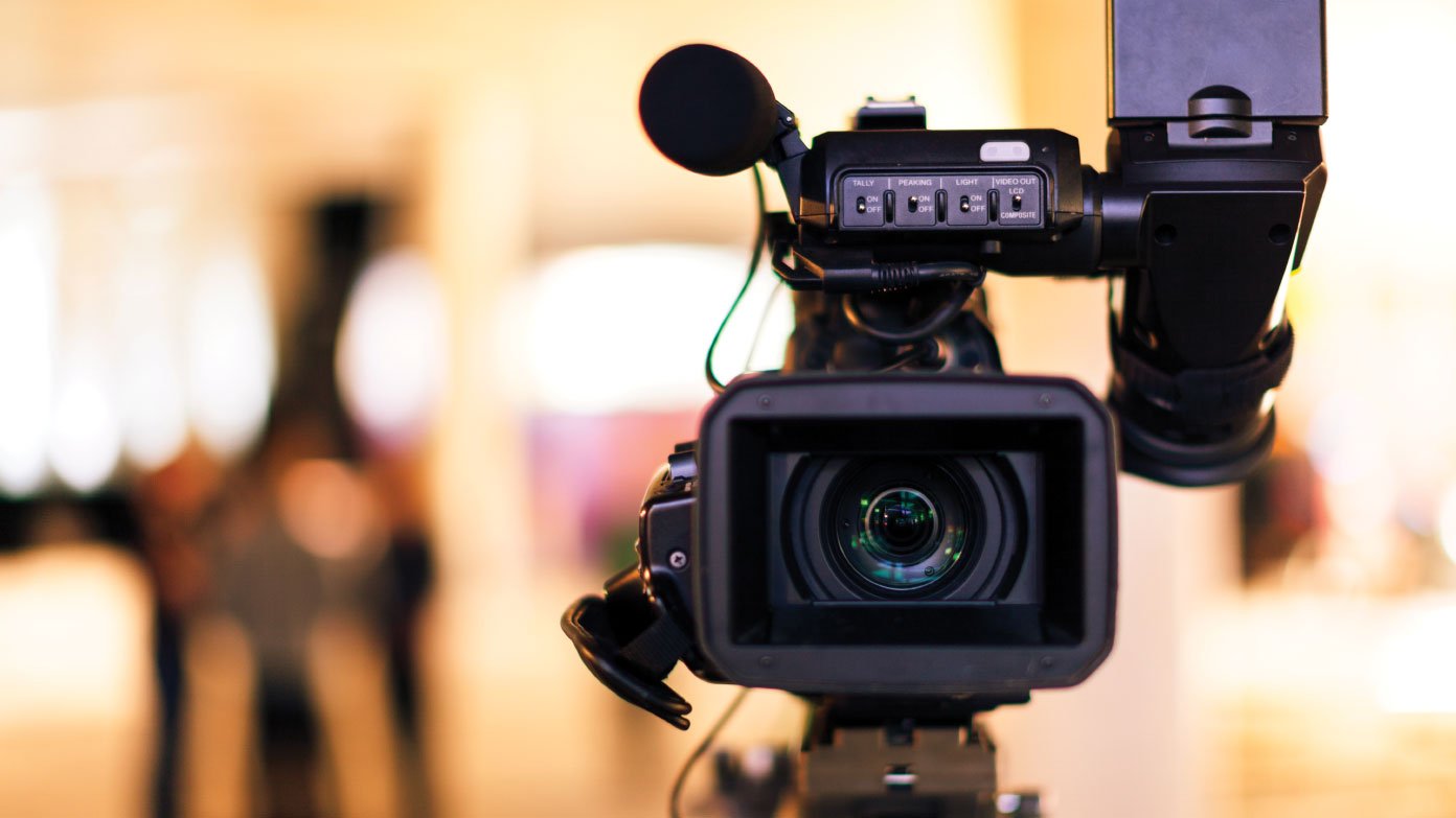 Close-up of a professional video camera with a large lens, set in a room with warm lighting and blurred background.