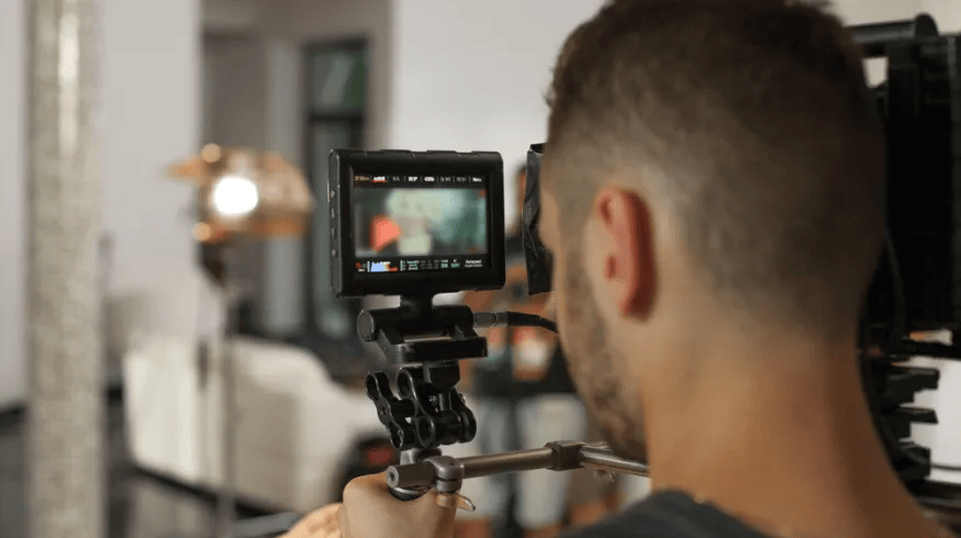 A person operating a professional video camera indoors, recording a scene with a blurred background of contemporary furniture.