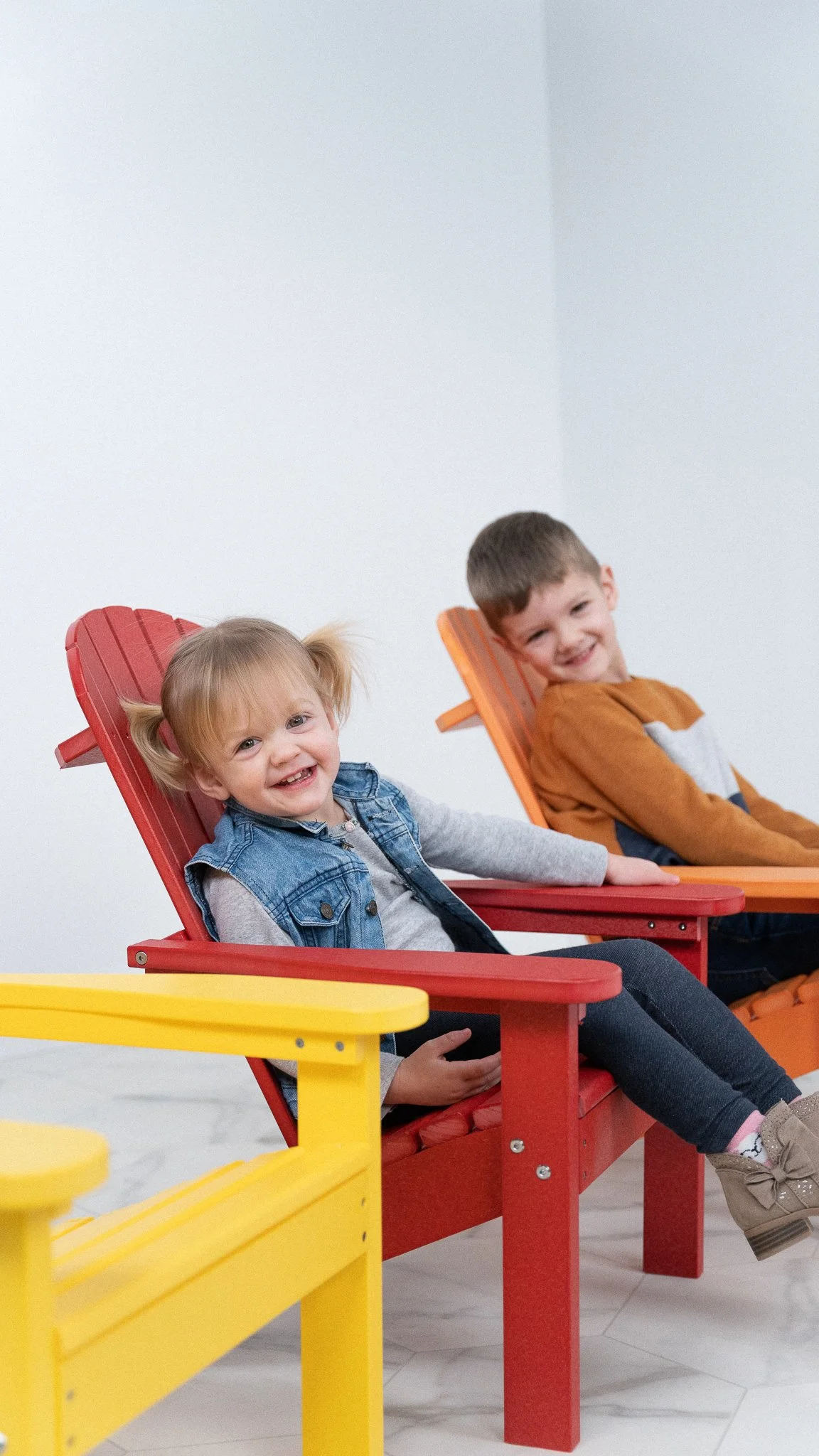 Two young children, a girl and a boy, sitting on colorful outdoor chairs indoors. The girl is smiling with pigtails, wearing a denim vest and grey long-sleeve shirt. The boy, in a brown and white sweater, is also smiling and leaning back on an orange