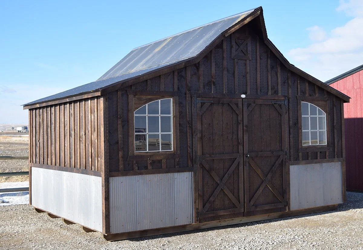 A small barn with dark wood siding, two windows, metal roof, and metal lower siding, situated on gravel ground in an outdoor setting.