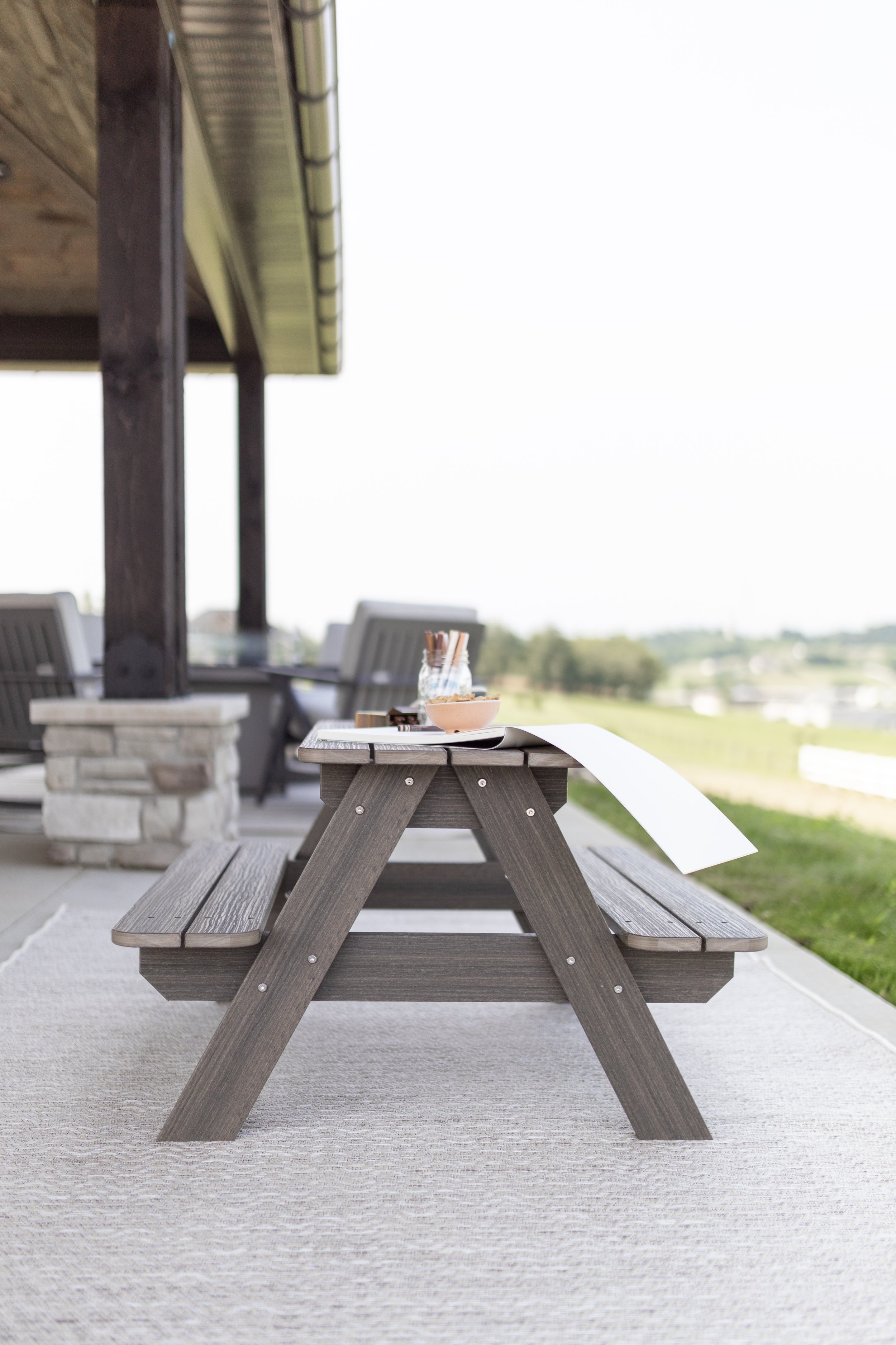 Wooden outdoor picnic table on a beige rug, with a bowl, a glass jar, and papers on top, overlooking a green landscape.