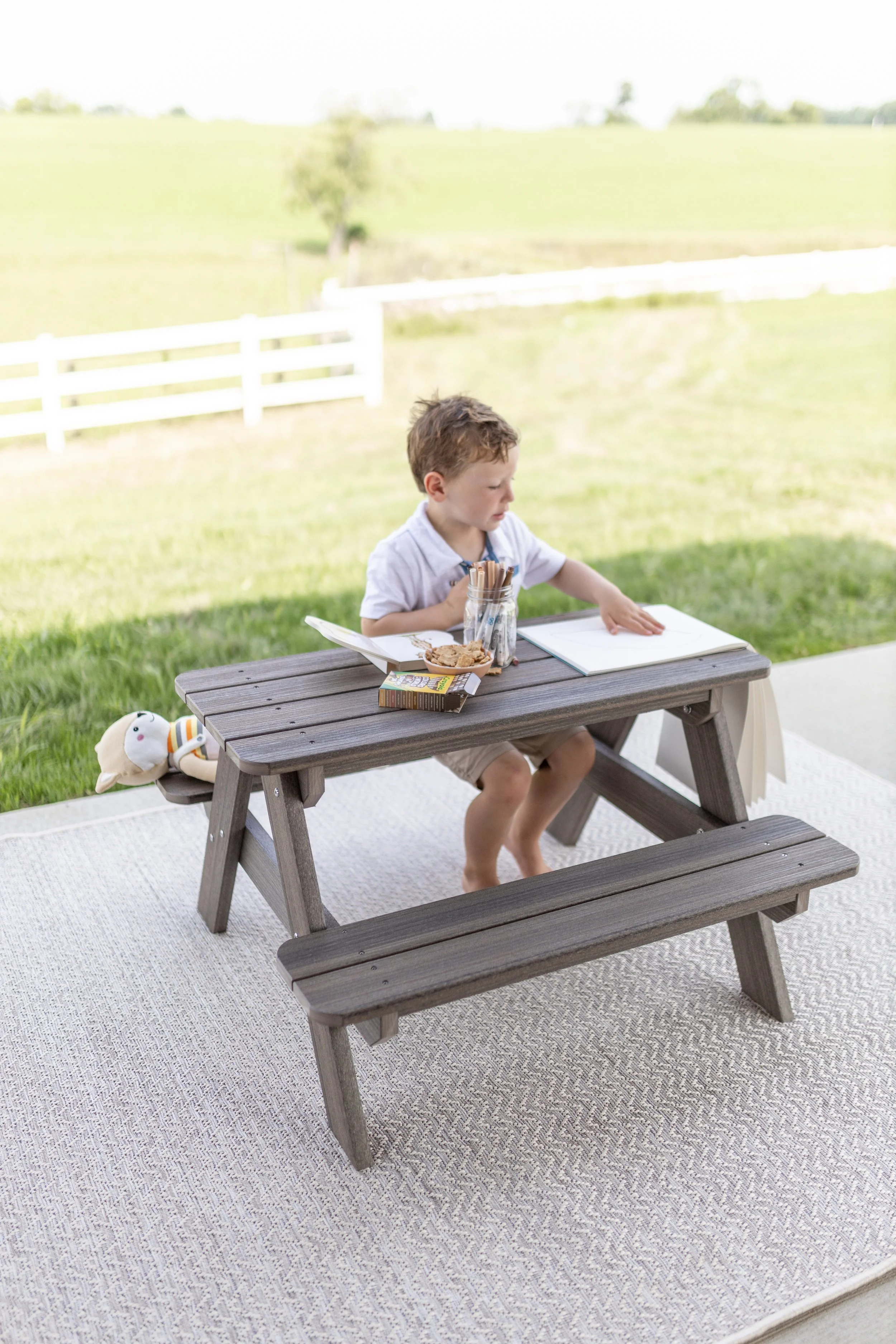 A young boy sitting at a wooden picnic table outside on a beige outdoor rug, with a white fence and green fields in the background. The boy is reaching for something on a blank piece of paper, with a jar of cookies and other snacks on the table.