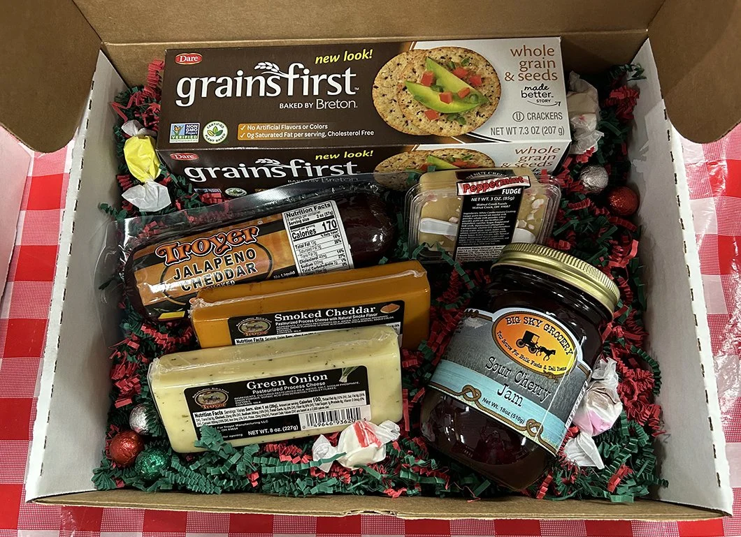 A gift box containing two boxes of whole grain and seeds crackers, a stick of Trojan jalapeño cheddar cheese, smoked cheddar cheese, green onion cheese, sour cherry jam, and assorted candies, all lined with red and green shredded paper on a red checkered tablecloth.