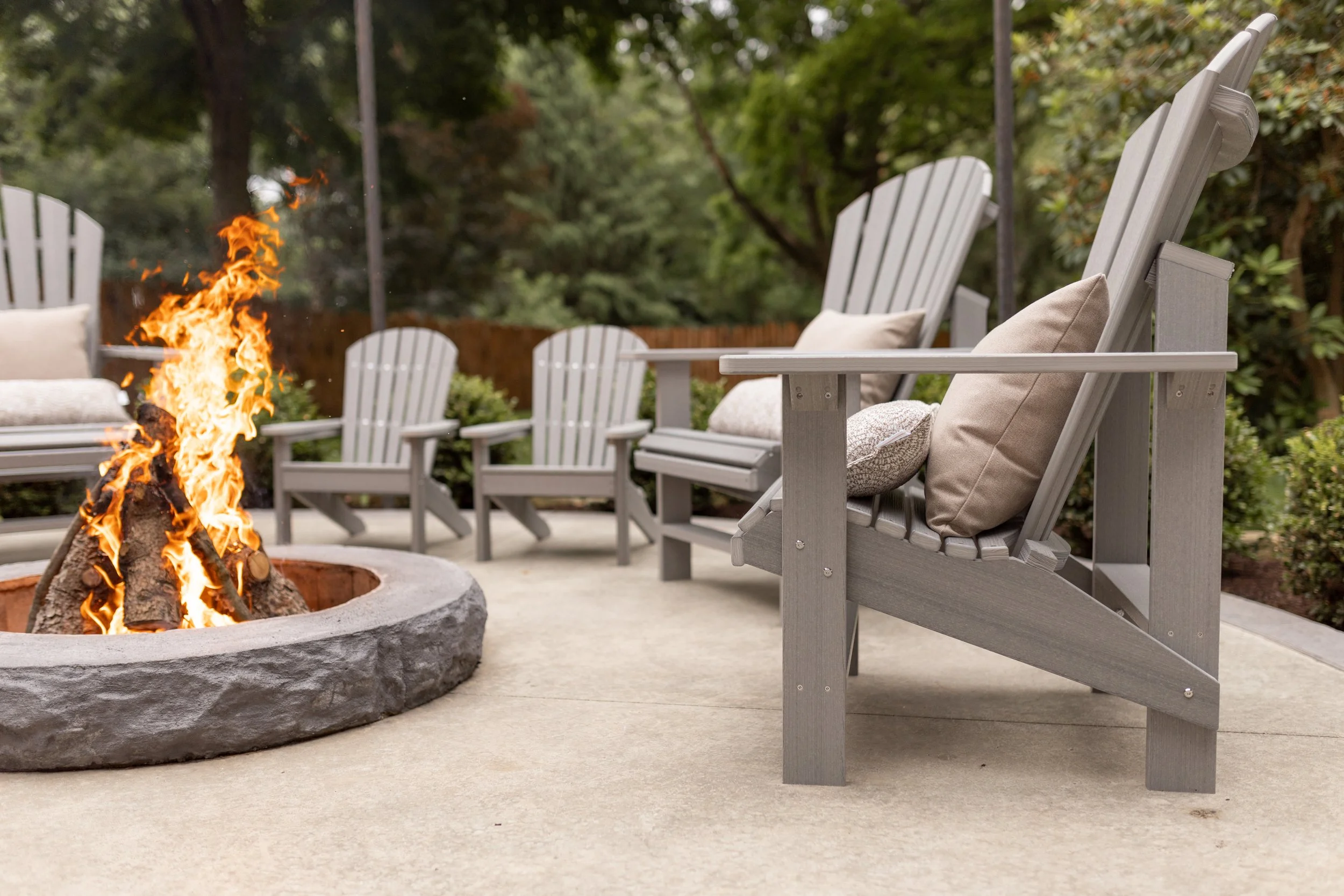 Outdoor patio with four gray Adirondack chairs arranged around a fire pit with a burning fire, surrounded by green trees and bushes.