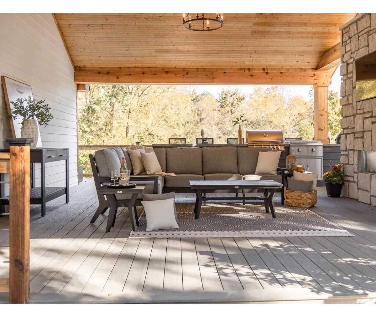 Outdoor patio with a large gray sectional sofa, a wooden coffee table, and side table on a patterned rug. There's a black grill and a counter with potted plants and candles. The space has a wooden ceiling and partial stone wall, with trees visible in the background.
