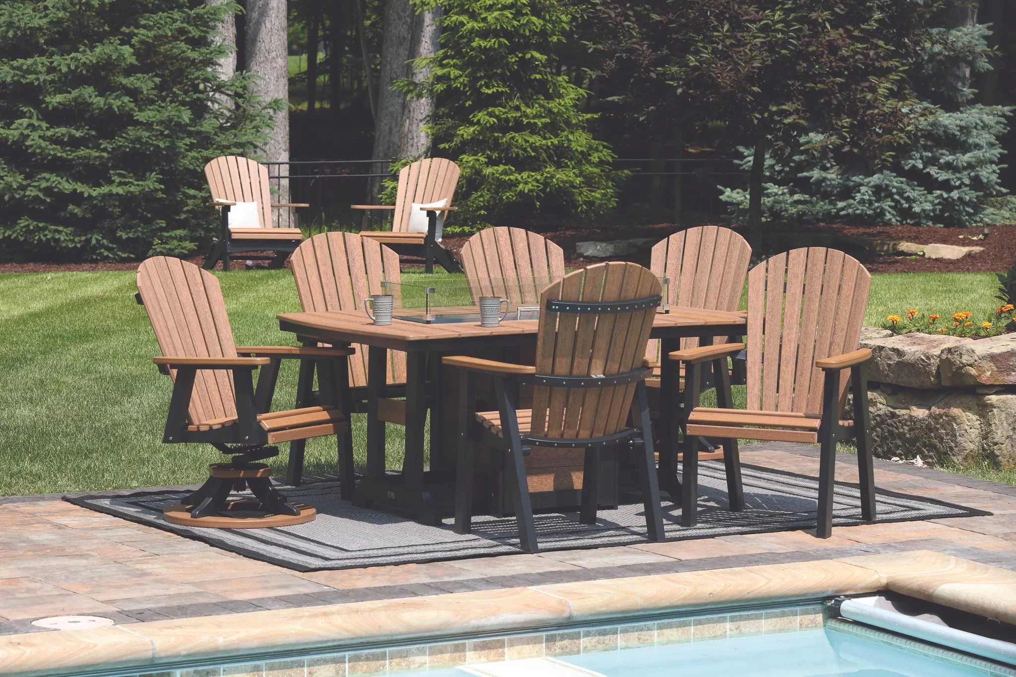 Outdoor patio area with a rectangular wooden dining table surrounded by wooden chairs, and two Adirondack chairs on the lawn in the background, with trees and shrubs.