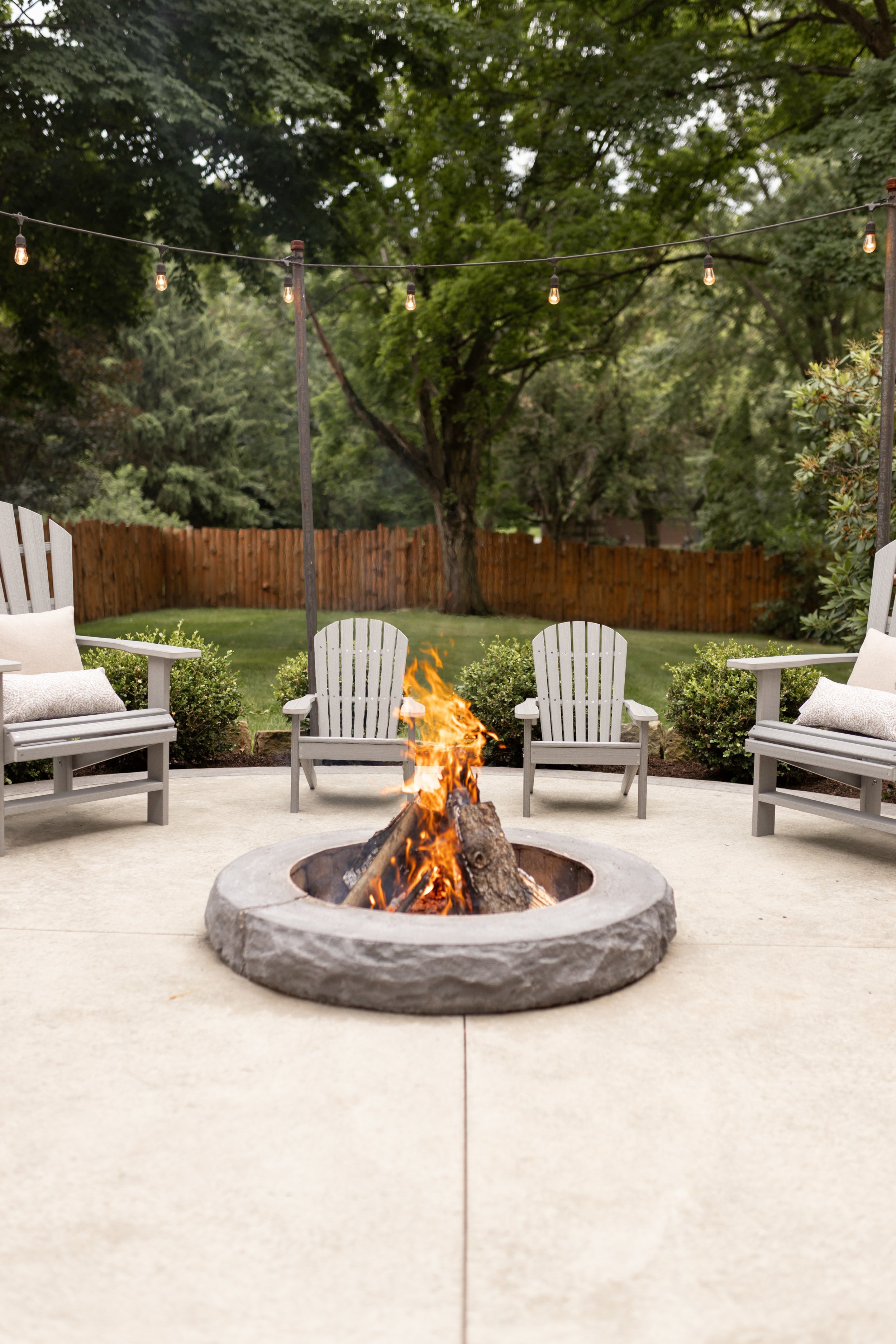 Backyard patio with a fire pit surrounded by four white Adirondack chairs and two white bench seats on either side. String lights hang above, and there is a wooden fence and trees in the background.