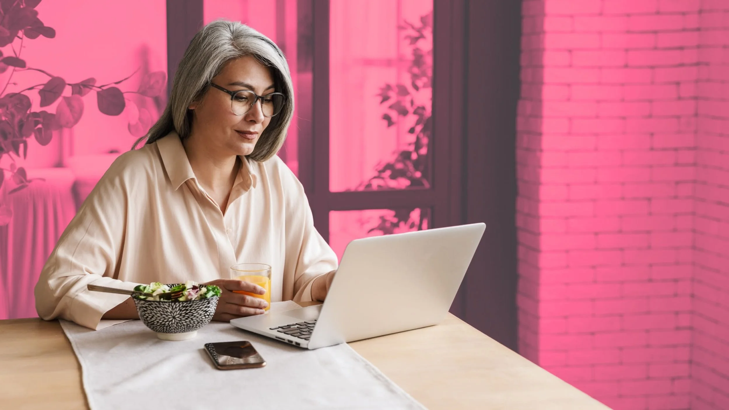 A woman with gray hair, glasses, and a beige blouse sitting at a wooden table, working on a laptop, with a bowl of salad and a glass of orange juice beside her, in a room with pink tinted windows and a brick wall.