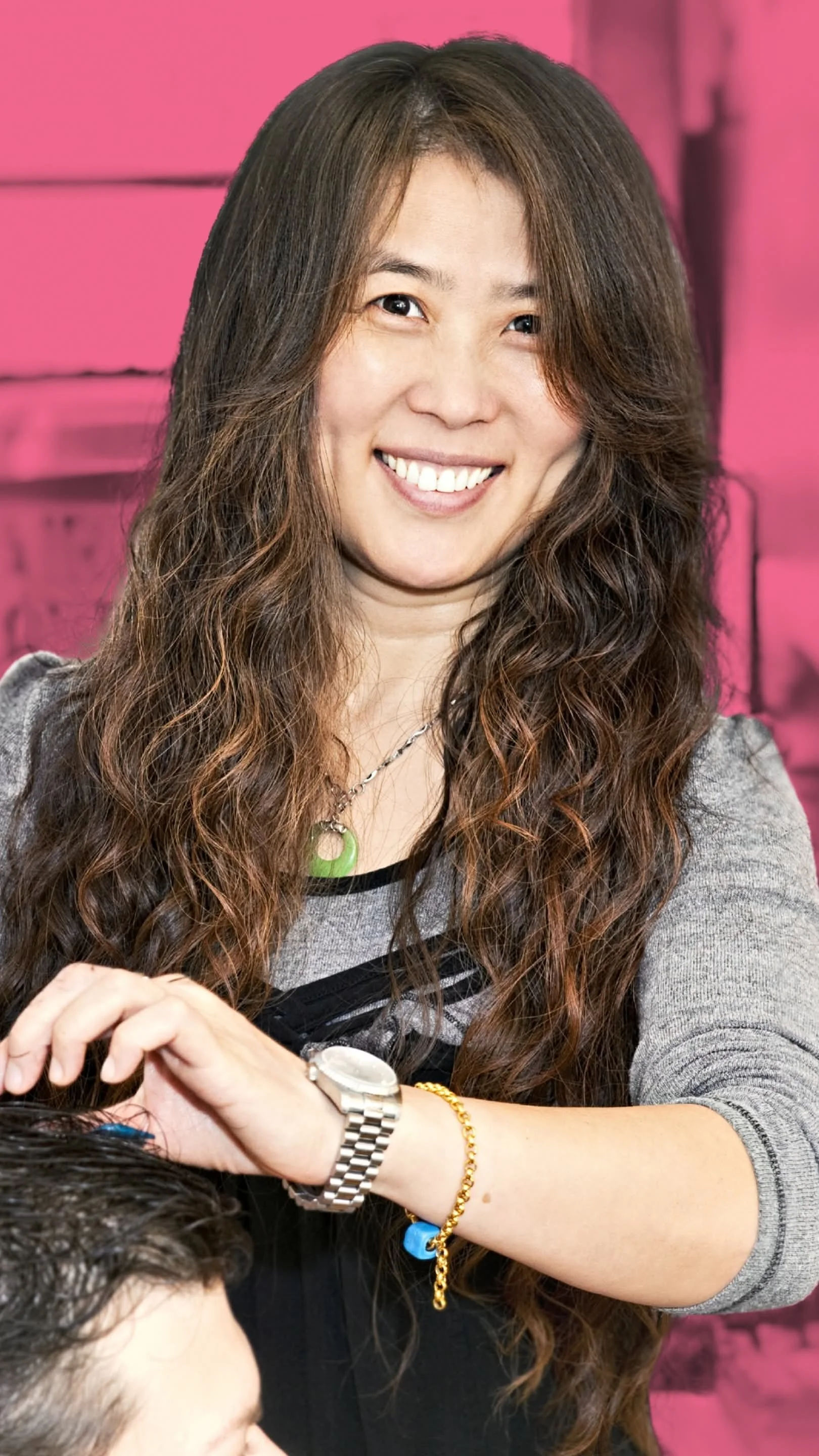 A smiling woman with long, curly brown hair, wearing a gray and black top, a silver watch, a gold chain bracelet, and a green necklace, appears to be styling someone's hair.