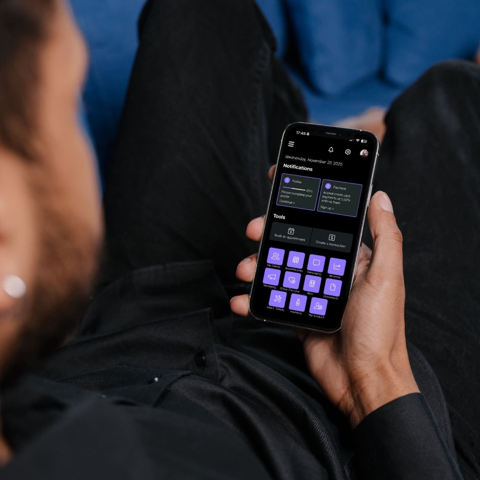 A person holding a smartphone in their hand, displaying a mobile app with notifications and icons on a dark background, while sitting with legs crossed and wearing dark clothing.