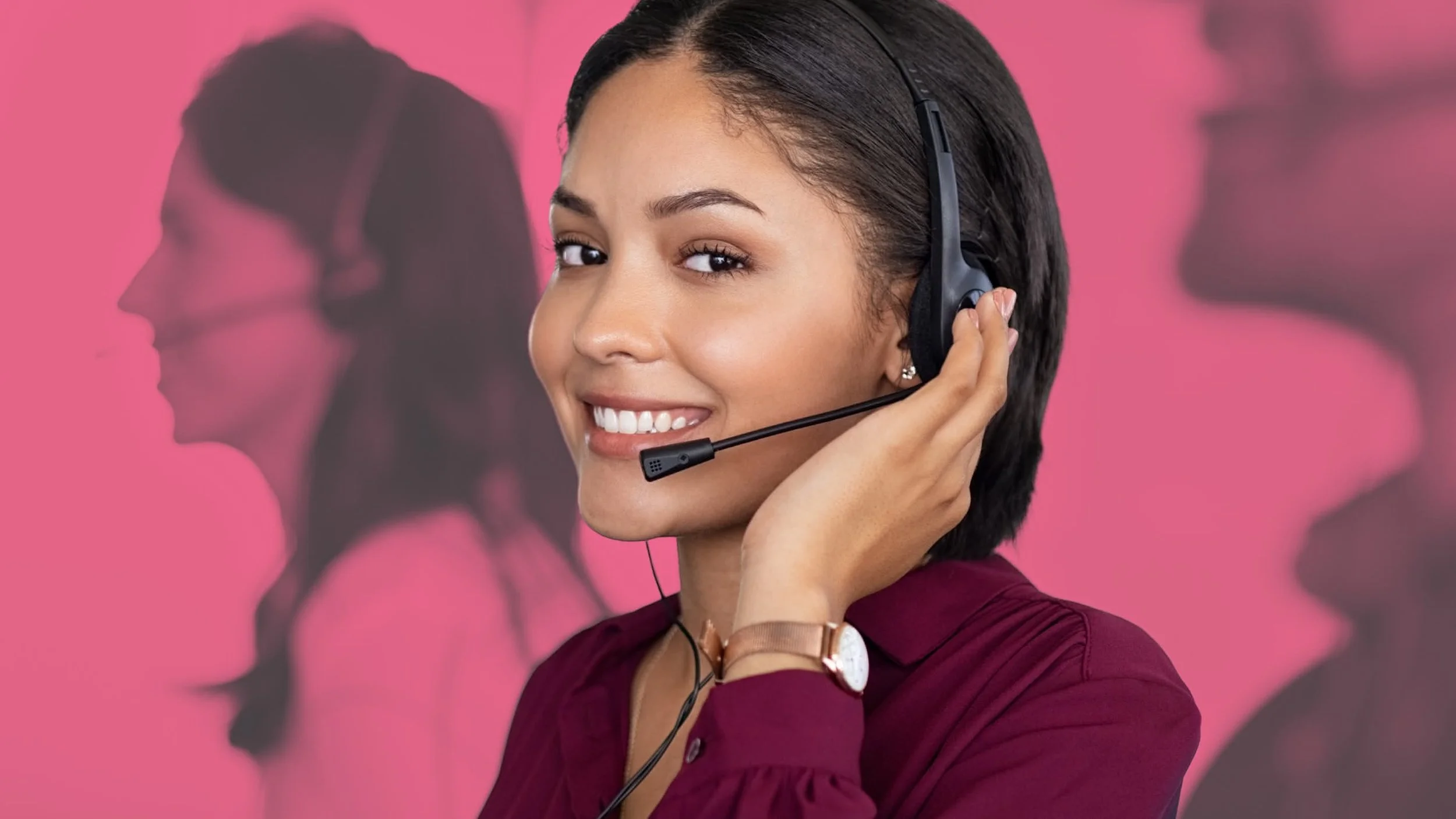 A smiling woman in a maroon top wearing a headset with a microphone, standing in front of a pink background with her reflection visible.